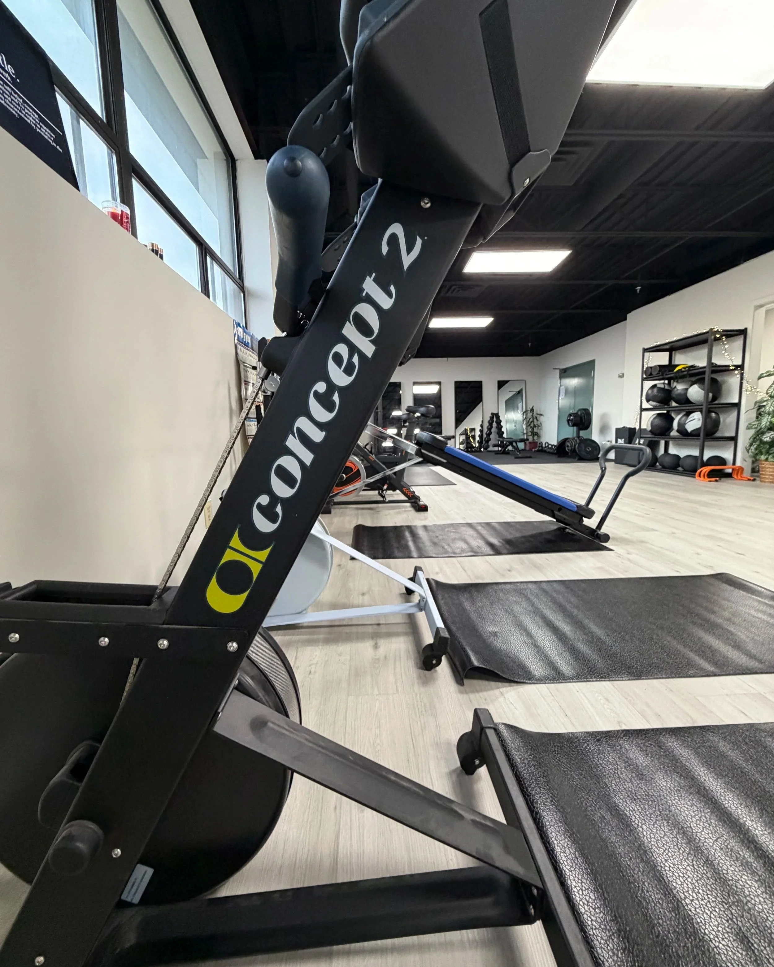 Interior of a fitness gym with exercise equipment, including a Concept2 rowing machine, yoga mats, and various weights on shelves.