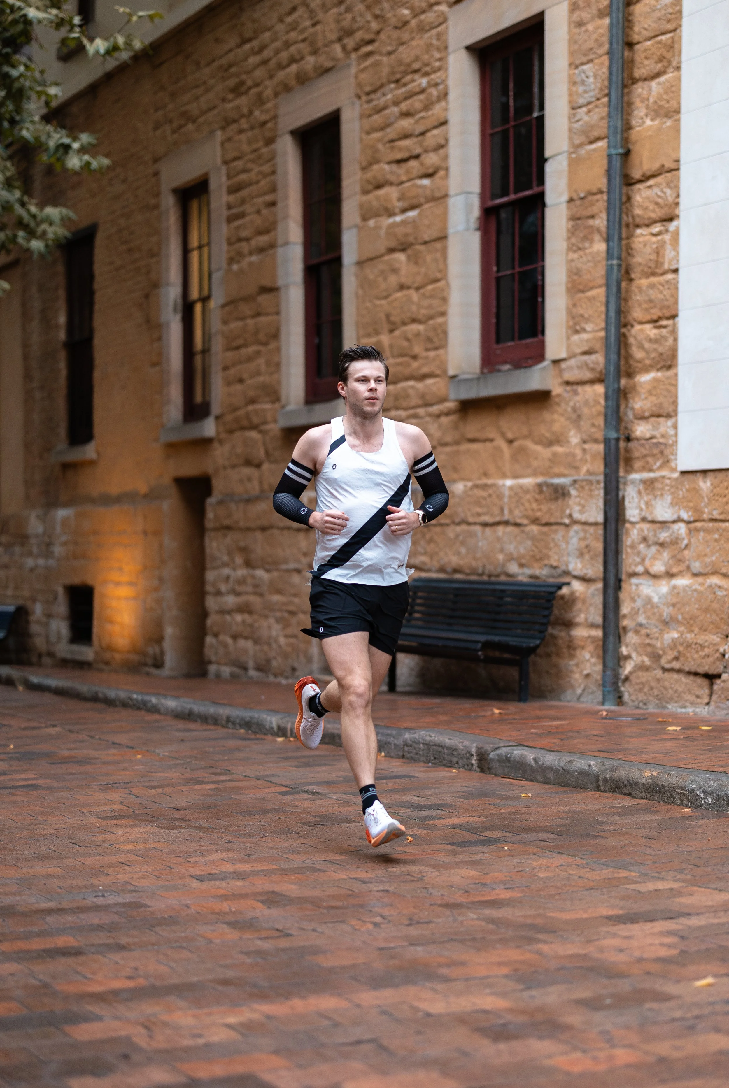 A man running on a brick sidewalk in front of a building with a stone facade and tall windows.