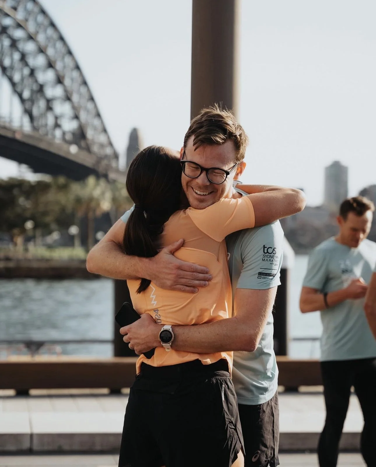 Two people hugging at a waterfront with a bridge in the background, one man smiling and wearing glasses, a woman with long dark hair, and a third person in the background jogging.