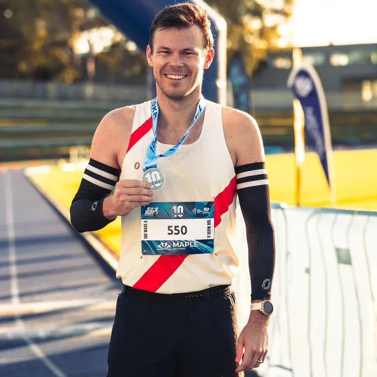 A smiling male marathon runner holding a silver medal, wearing a white and red running singlet, black arm sleeves, a race bib with the number 550, and a watch, standing on a race track during sunset.