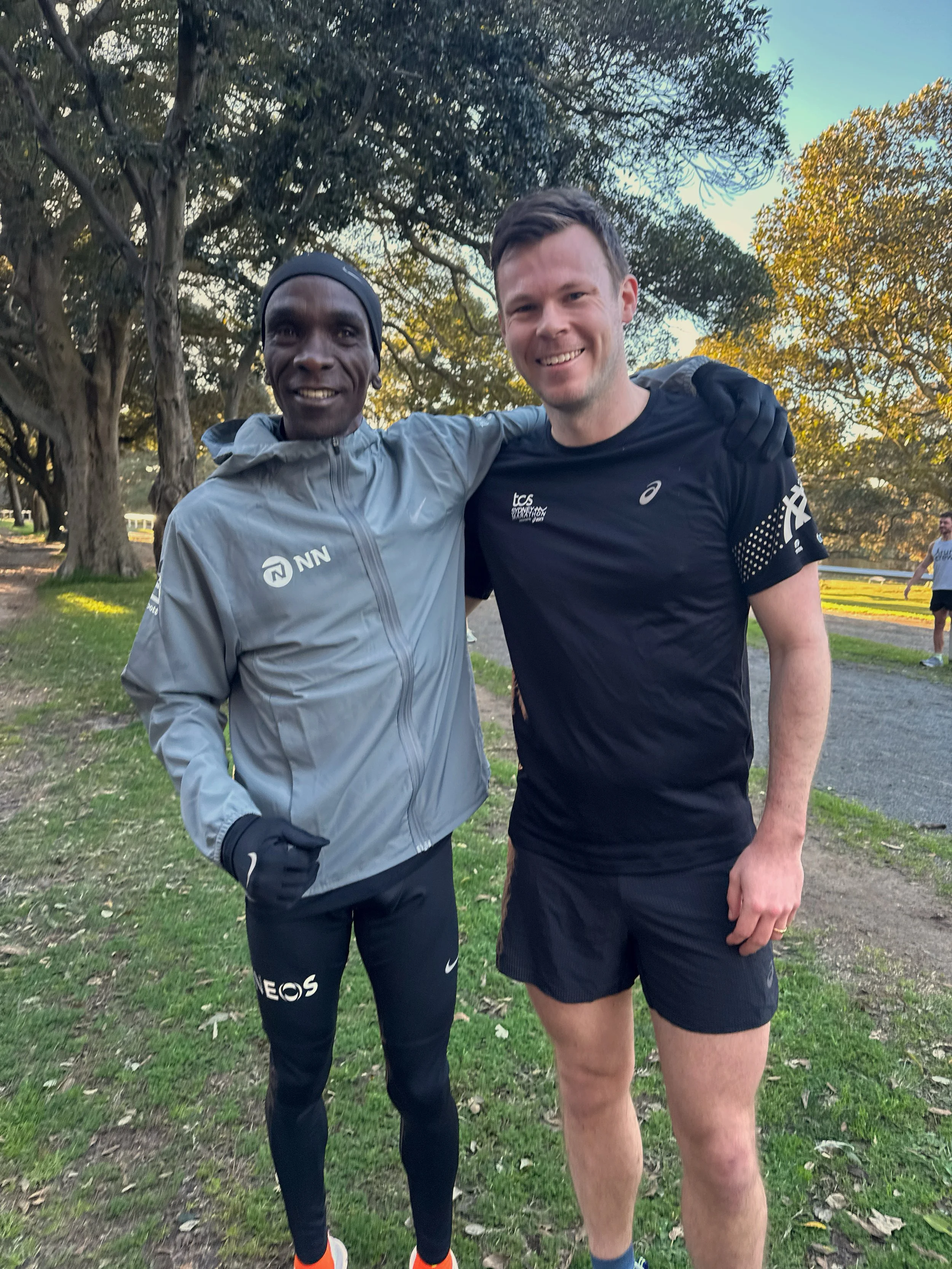 Two men standing outdoors on a grassy area, smiling with their arms around each other. The man on the left is wearing a gray jacket, black pants, gloves, and a headband, and the man on the right is shirtless, wearing black shorts and a black shirt. There are large trees and blue sky in the background.