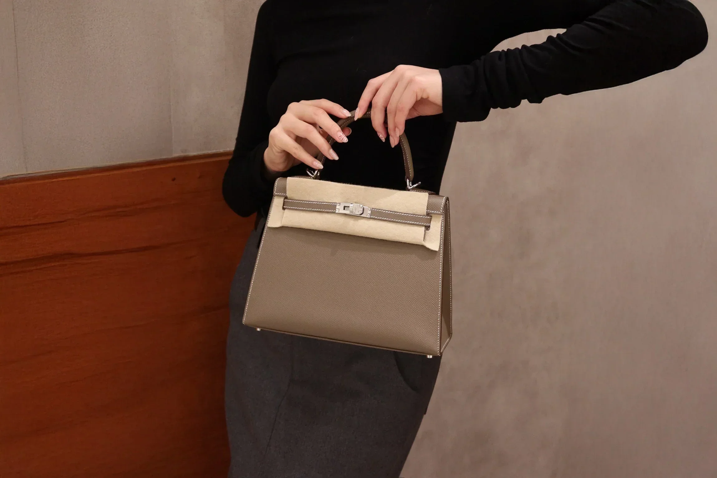 Person wearing black top holding a beige and brown handbag with a top handle, standing against a wooden wall and beige background.