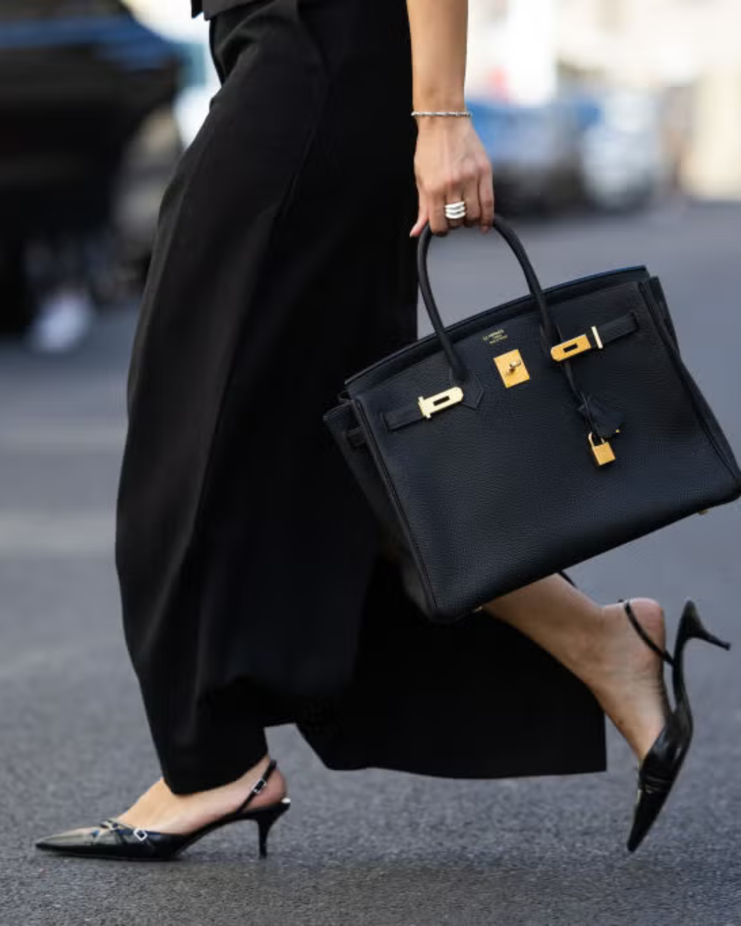 Birkin 30 black togo with gold hardware.  A woman dressed in black high heels and black pants walking on street while holding a black designer handbag with gold hardware.