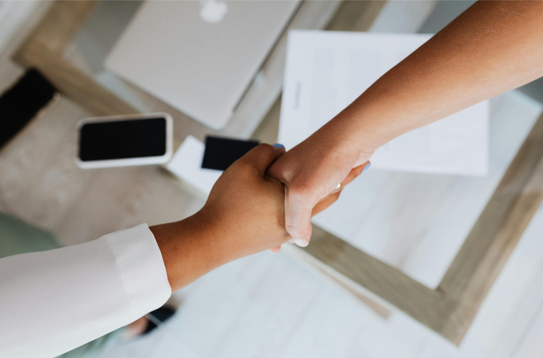 Two people shaking hands over a table with papers, a mobile phone, and a laptop.