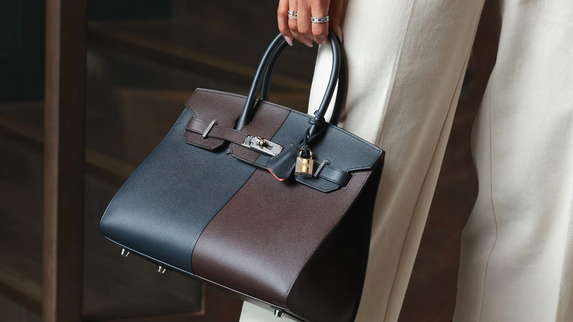 Birkin 30 sellier style A person holding a designer handbag with a lock, padlocks, and a strap attached, in black, brown, and navy colors, while wearing white pants and rings.