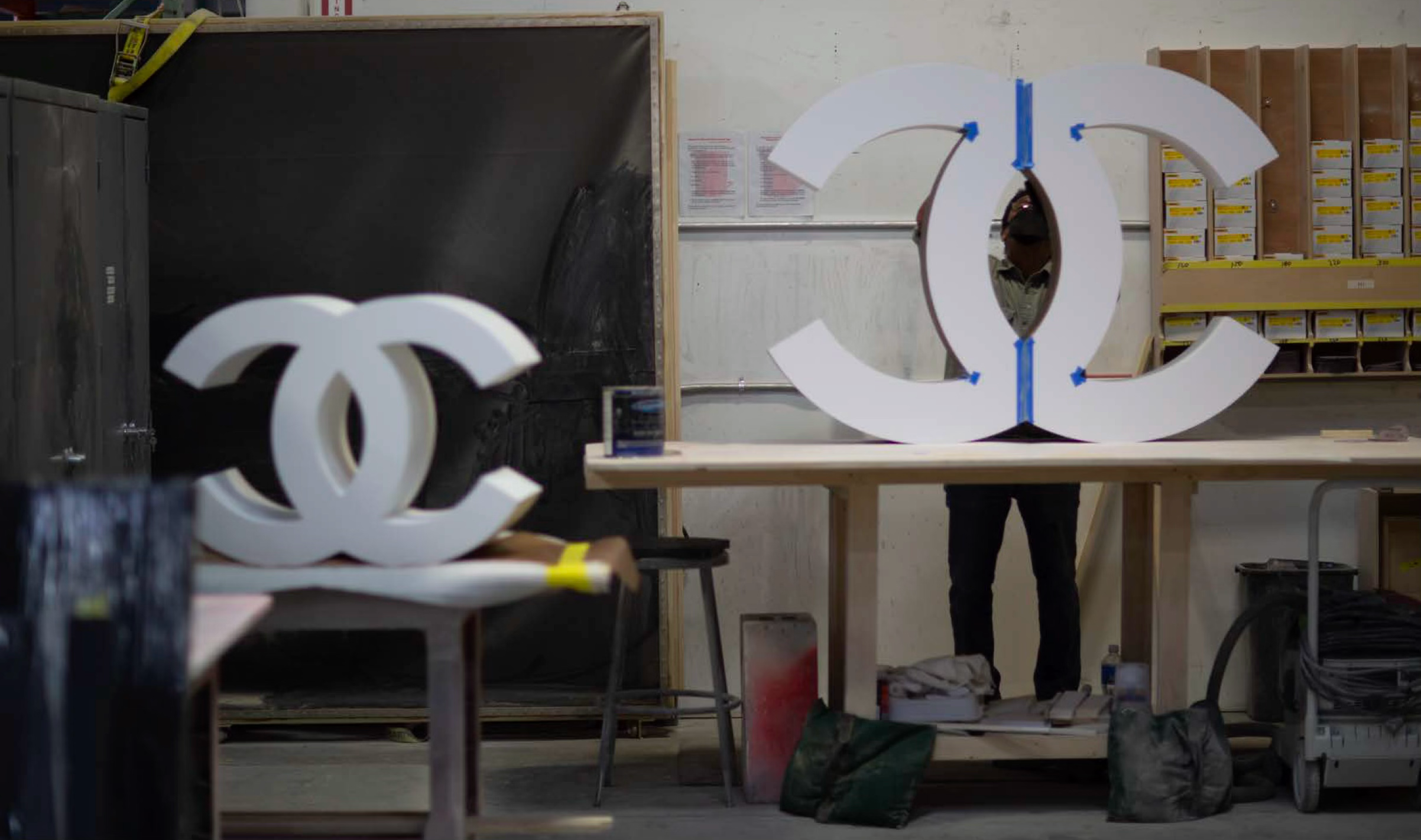 Workshop with large white decorative letter 'C' and 'O', possibly for a sign or display, set on a worktable. The workshop contains shelves with boxes, tools, and equipment, with a person partially visible behind the letter.
