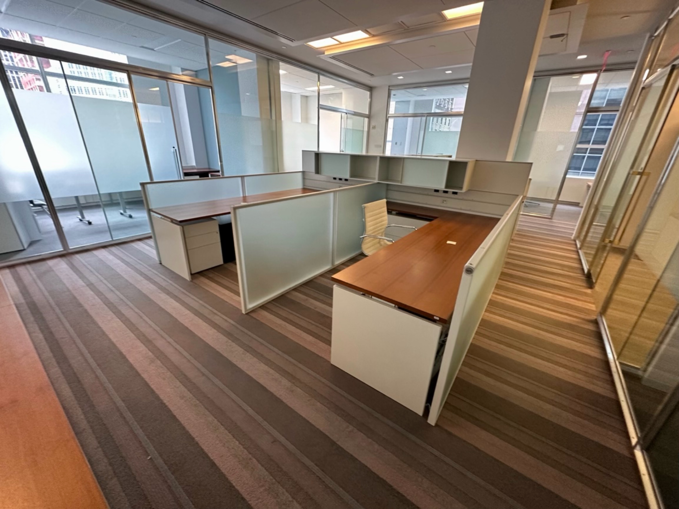 An office space with multiple workstations separated by low partitions, large windows with city buildings outside, and an open layout.