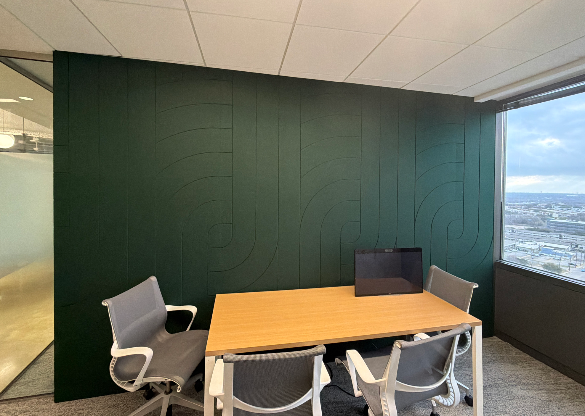 An office conference room with a rectangular wooden table, several white and gray chairs, a green accent wall with a geometric pattern, and floor-to-ceiling windows showing a cityscape and blue sky outside.