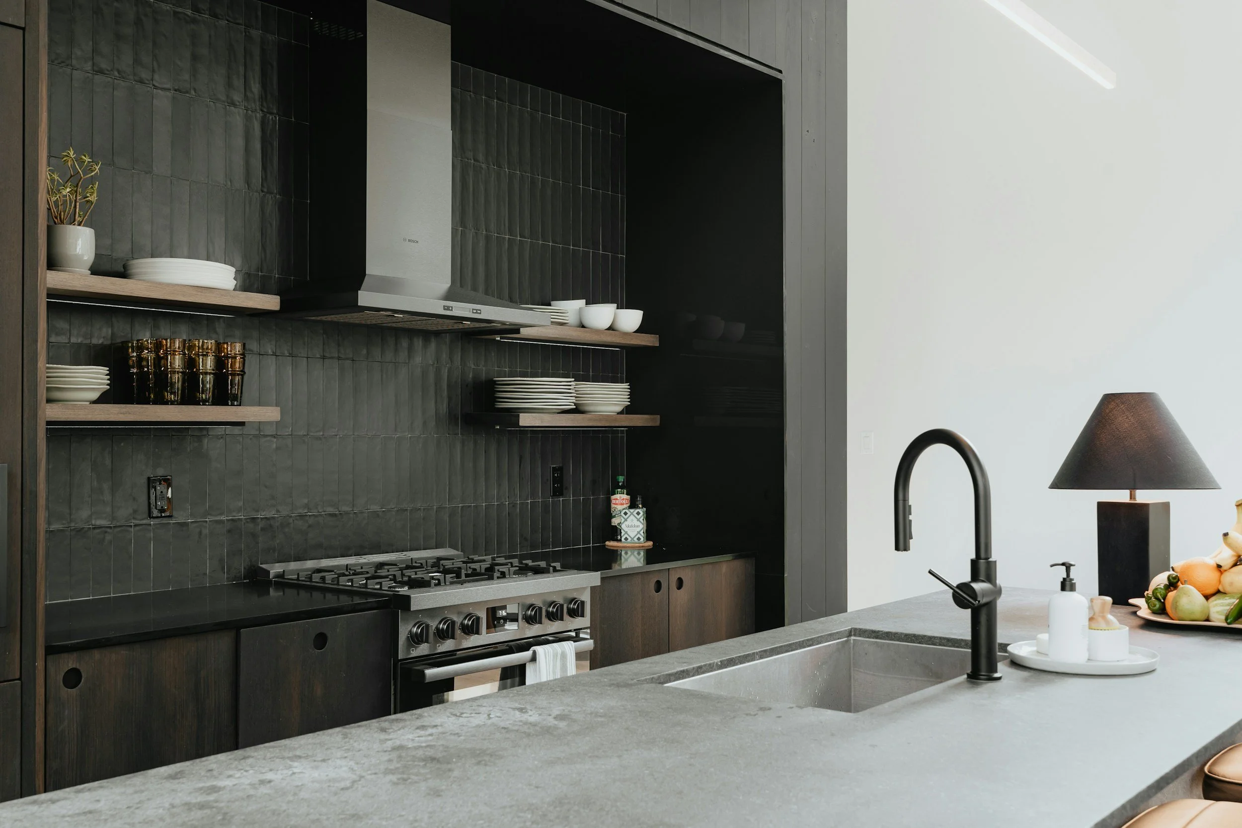 Modern kitchen with black tiled backsplash, open wooden shelves holding white dishes and glasses, stainless steel stove and range hood, a gray countertop with a black sink and faucet, and a black table lamp with a tray of fruits and toiletries.