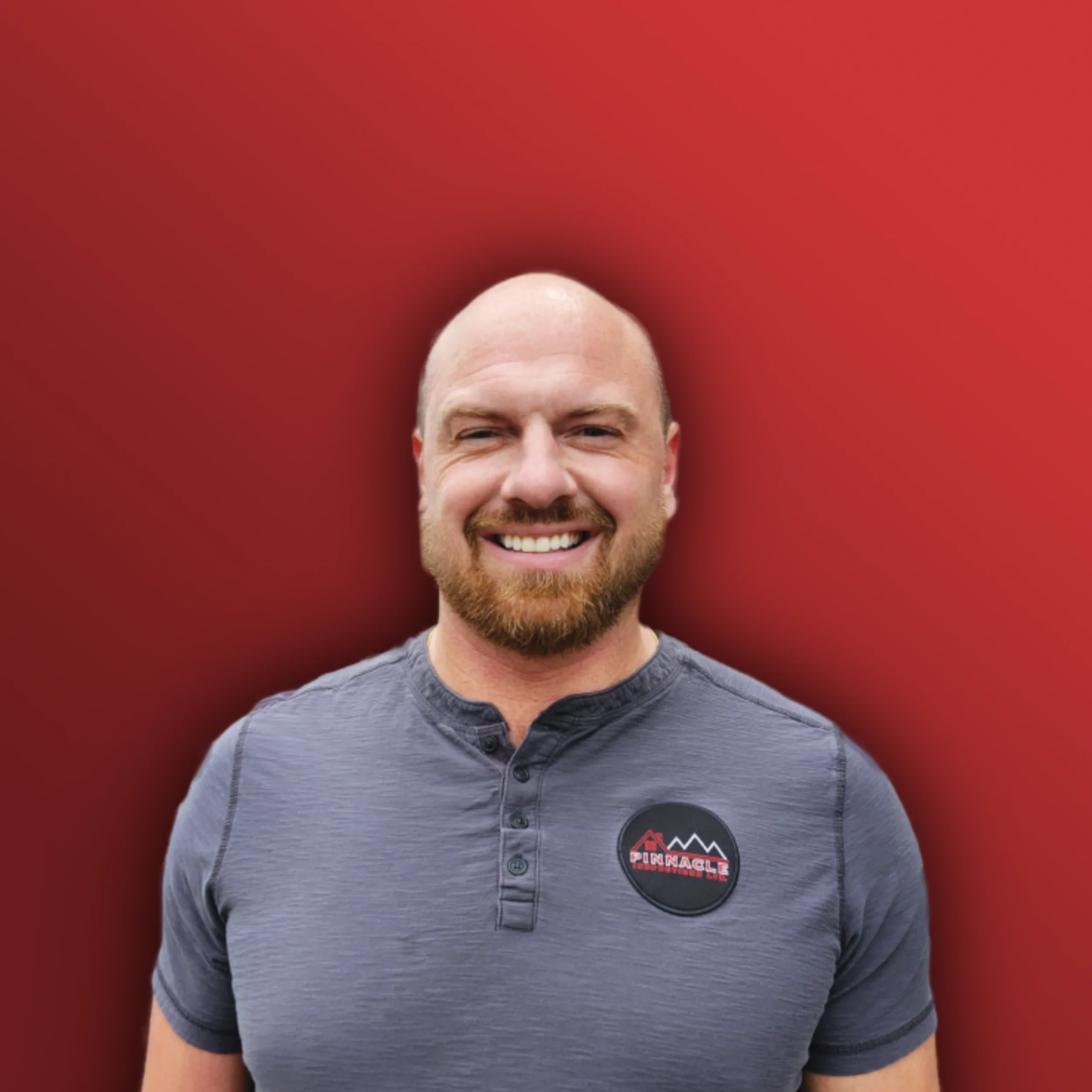 A smiling man with a shaved head, beard, and gray shirt standing against a red background.