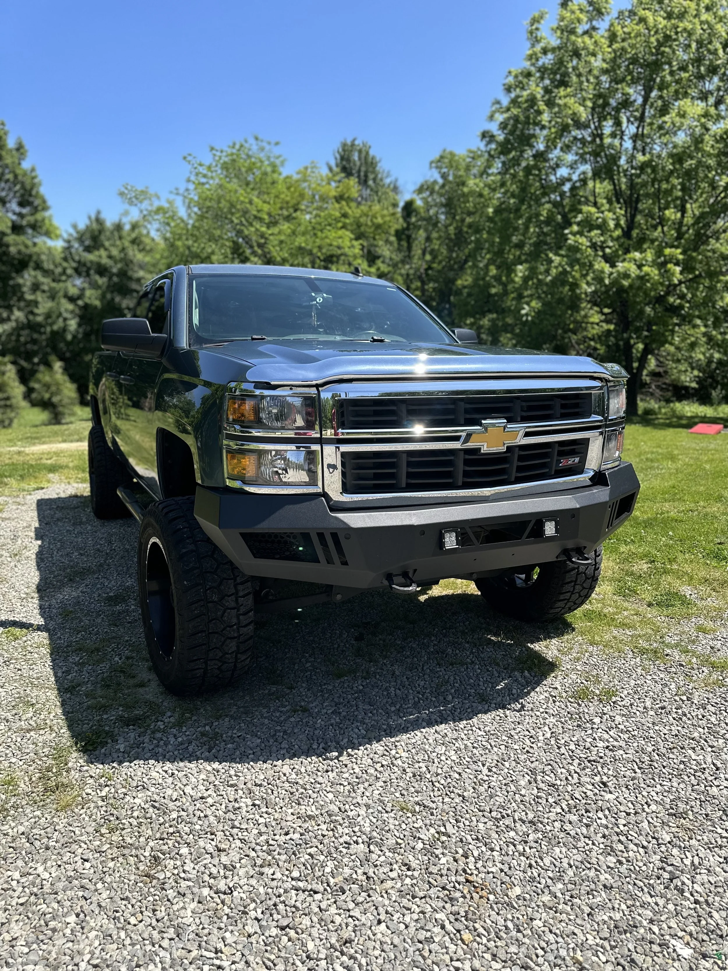 A black Chevrolet Silverado pickup truck parked on gravel, with trees and a clear blue sky in the background.