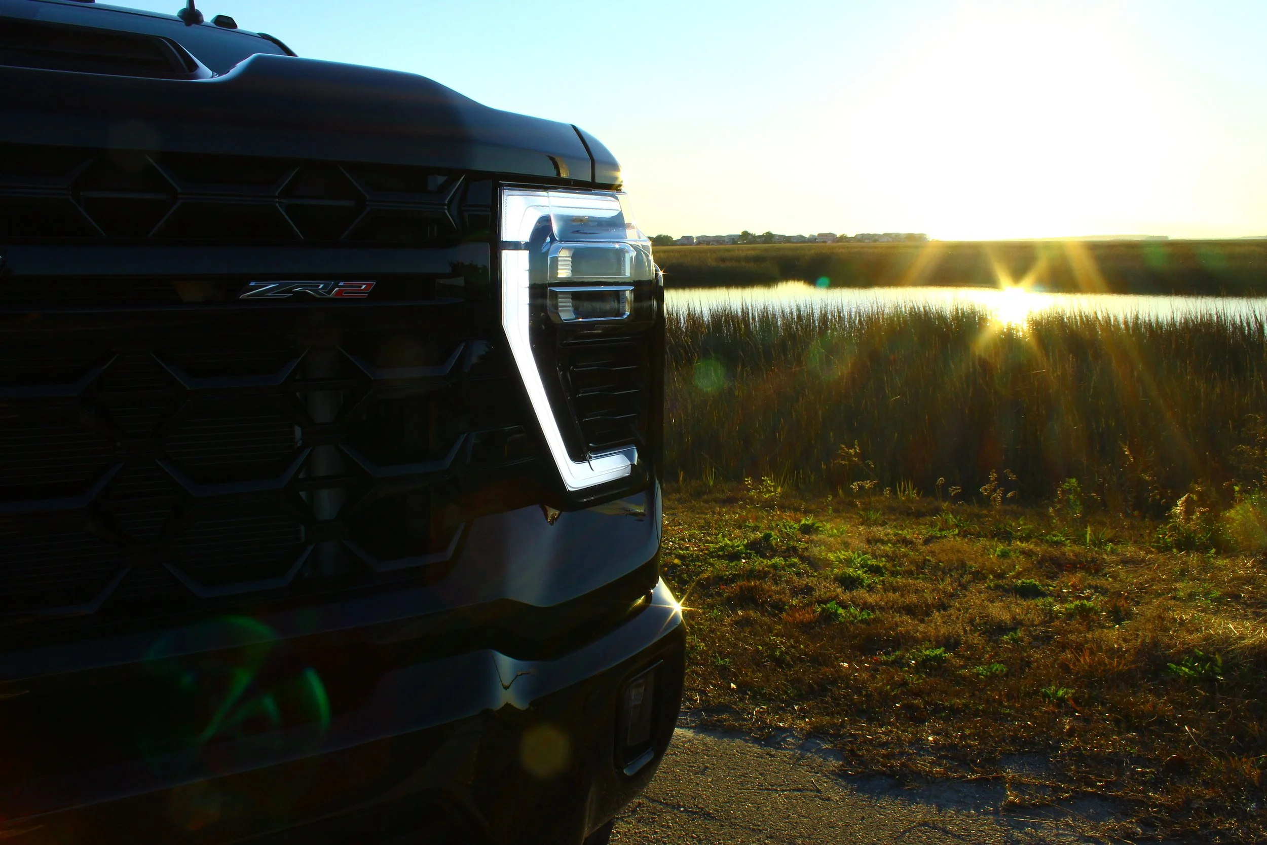 Close-up of the front left side of a black Chevrolet ZR2 pickup truck parked by a grassy field with a water body in the background, during sunset with the sun low on the horizon.