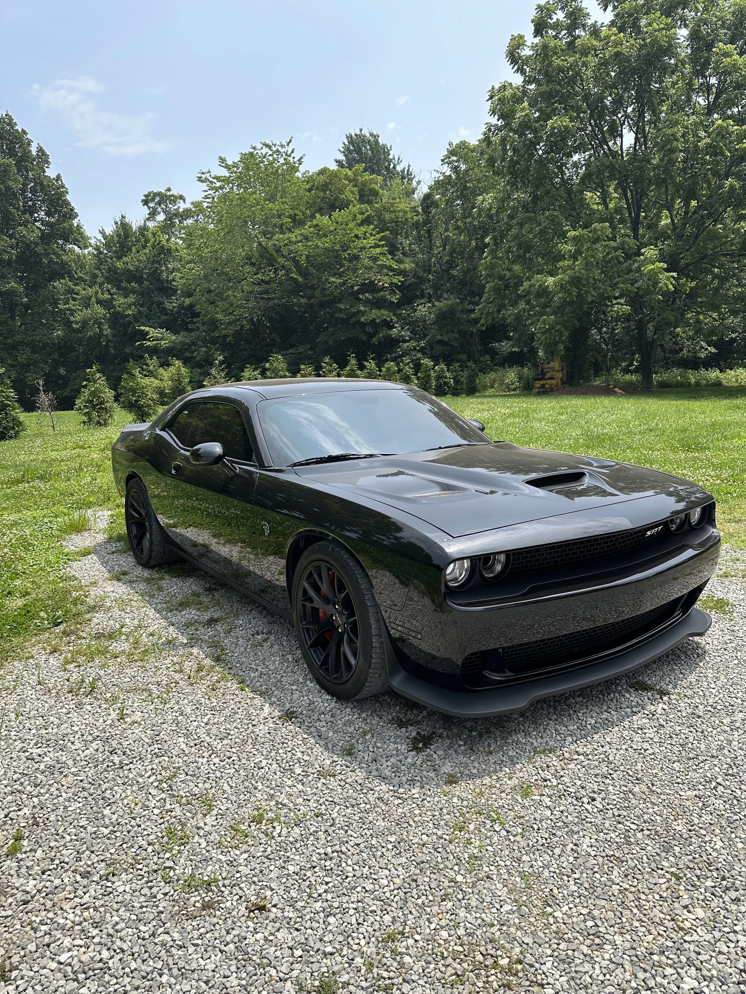 Black Dodge Challenger SRT parked on gravel with green trees and blue sky in the background.