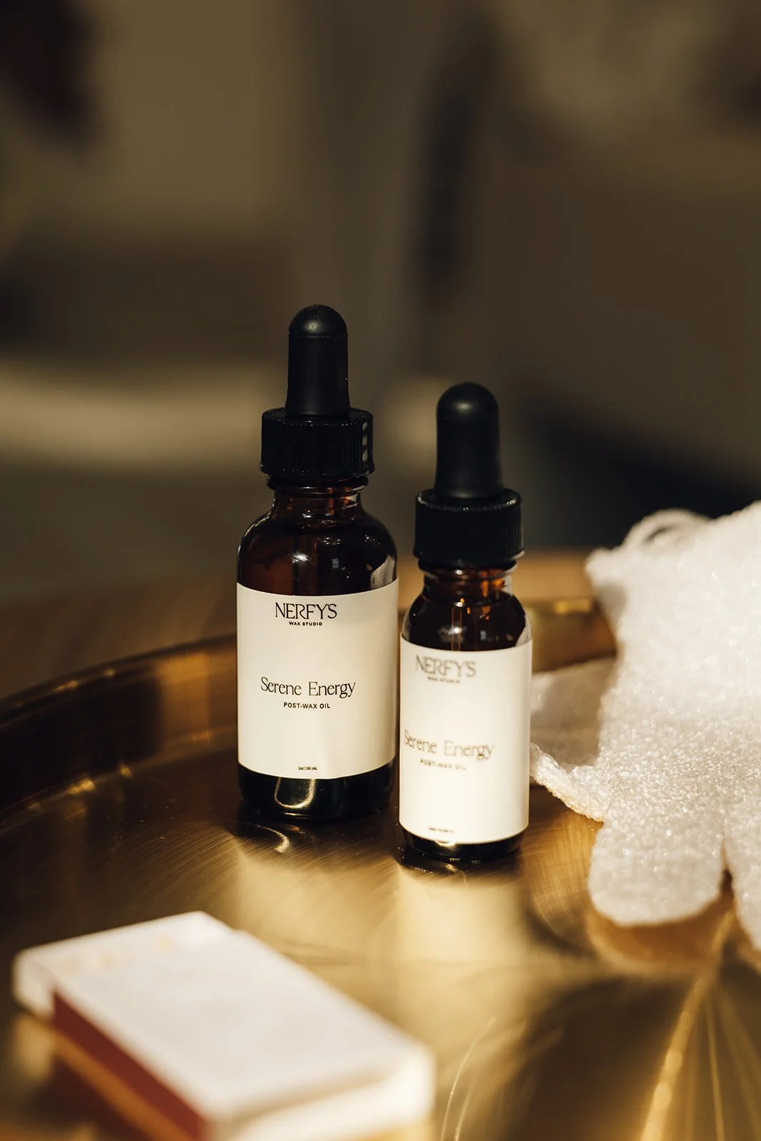 Two bottles labeled 'Serene Energy Post-Wax Oil' on a golden tray, with a white sponge nearby.