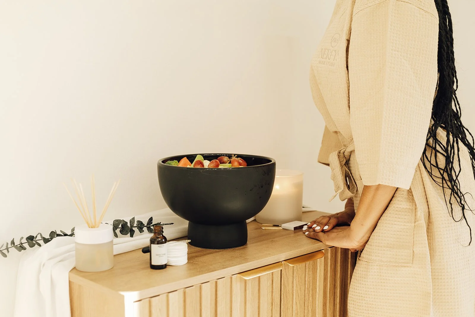 A woman standing at a wooden table with a black bowl of salad, a lit candle, facial oils, and a diffuser with reed sticks, in a minimalist setting.