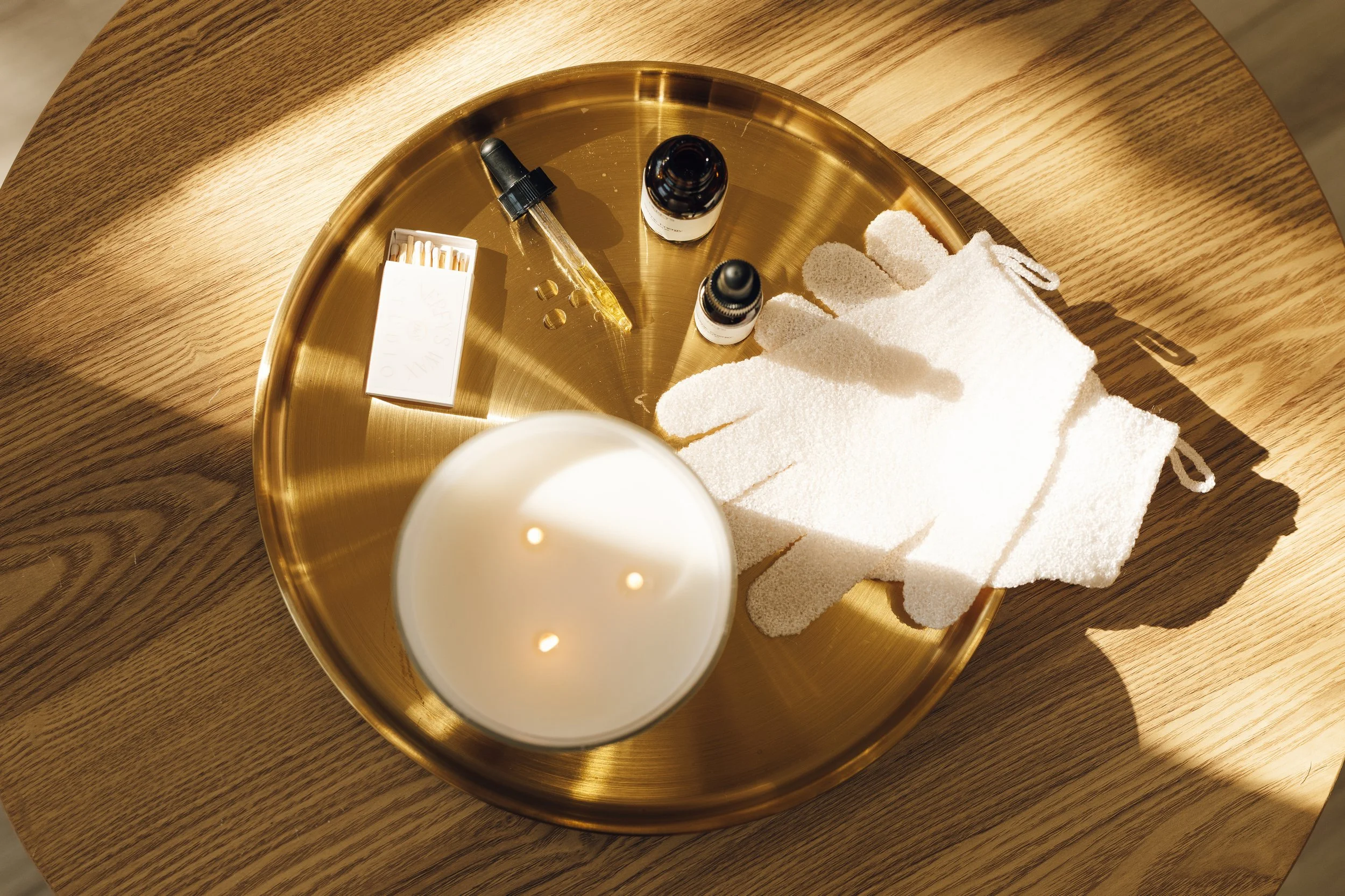 A round, gold-colored tray on a wooden table holding a white candle, two small bottles of essential oils, a glass dropper, a box of matches, and a pair of white spa gloves.