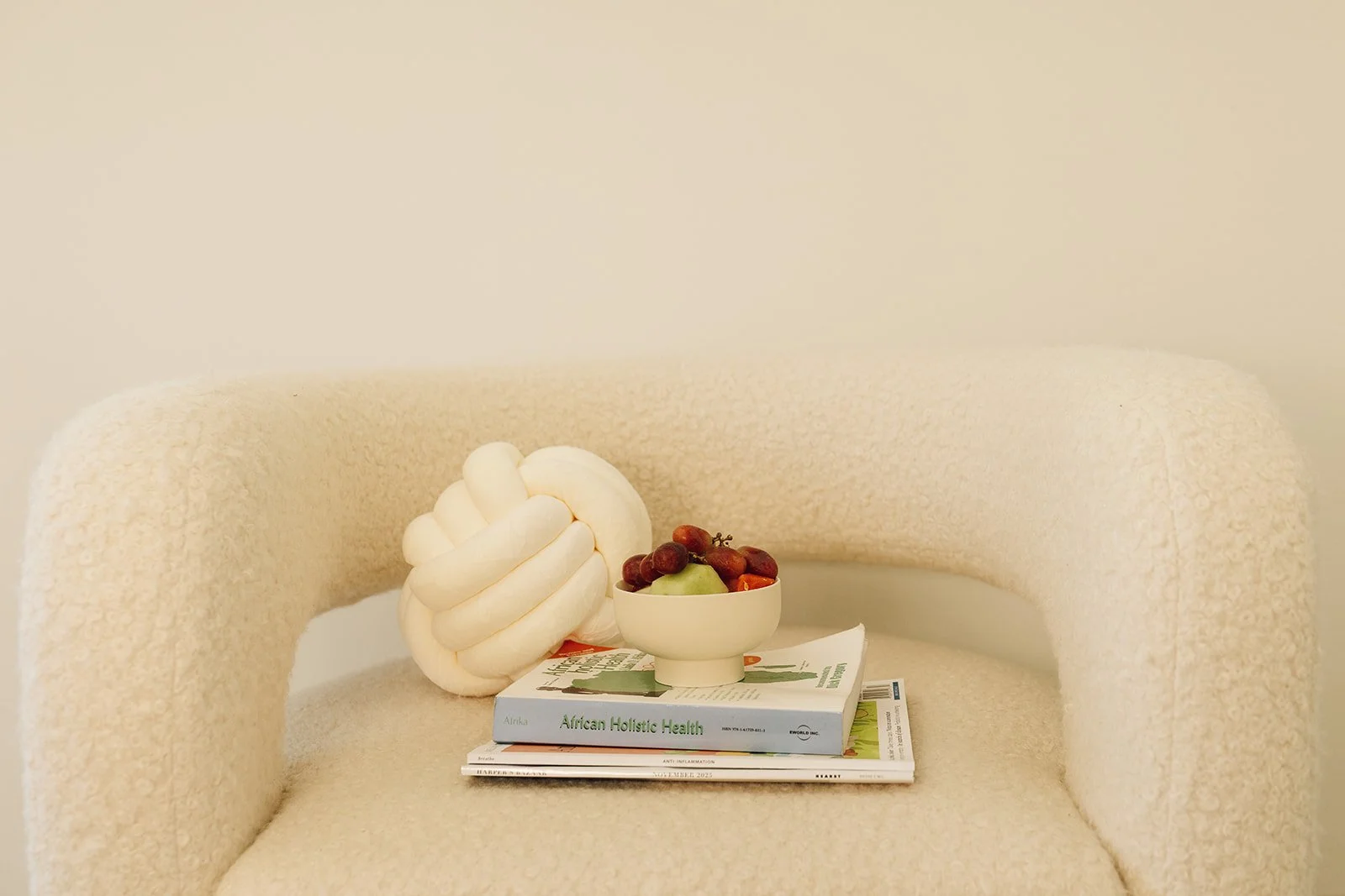 A beige textured armchair with a stack of magazines, a white bowl of assorted grapes, and a decorative white twisted rope knot on top of the magazines.