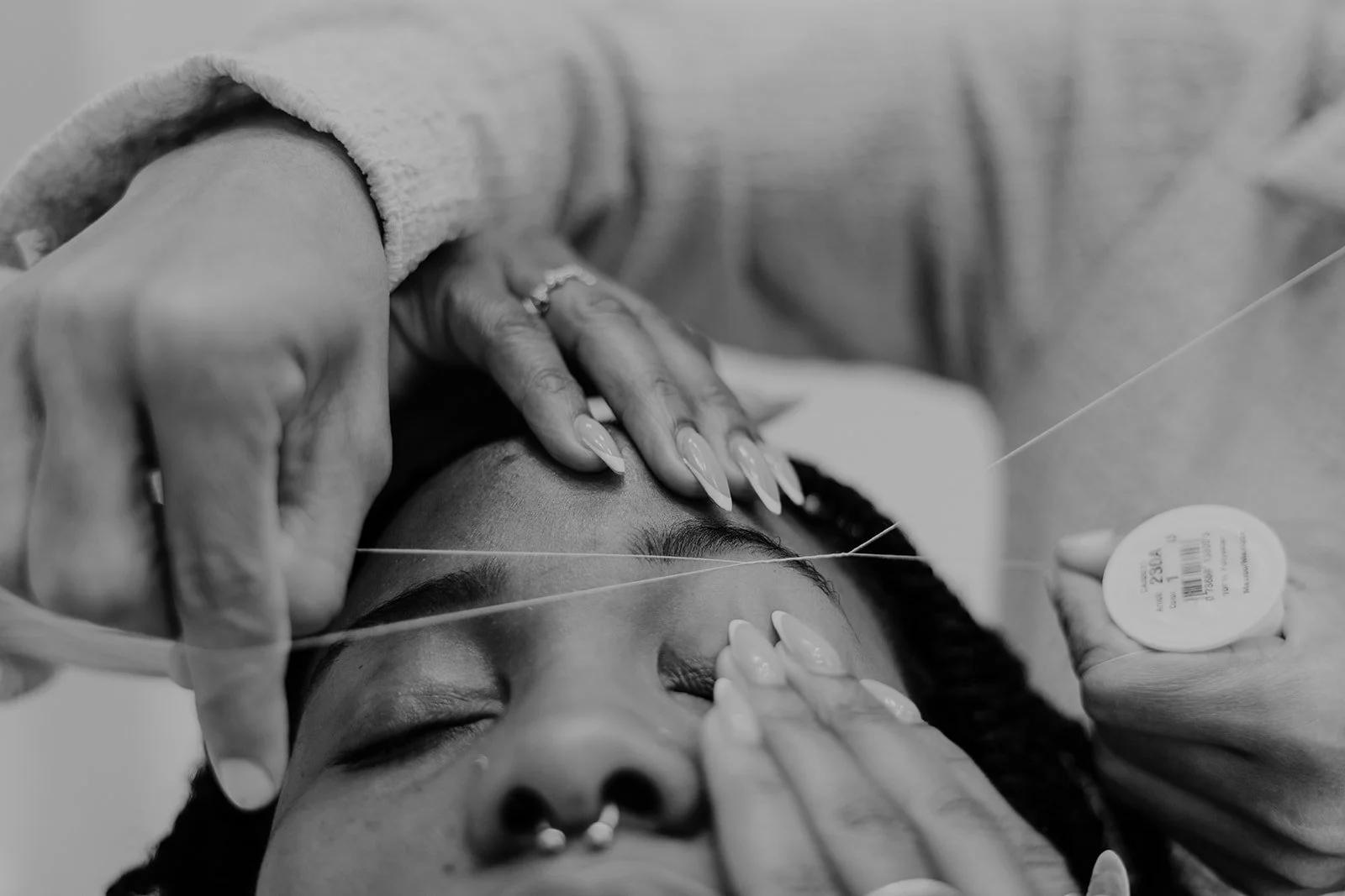 A person receiving a facial, threading, or skincare treatment, with hands carefully applying or adjusting over the face, in a close-up black and white photograph.
