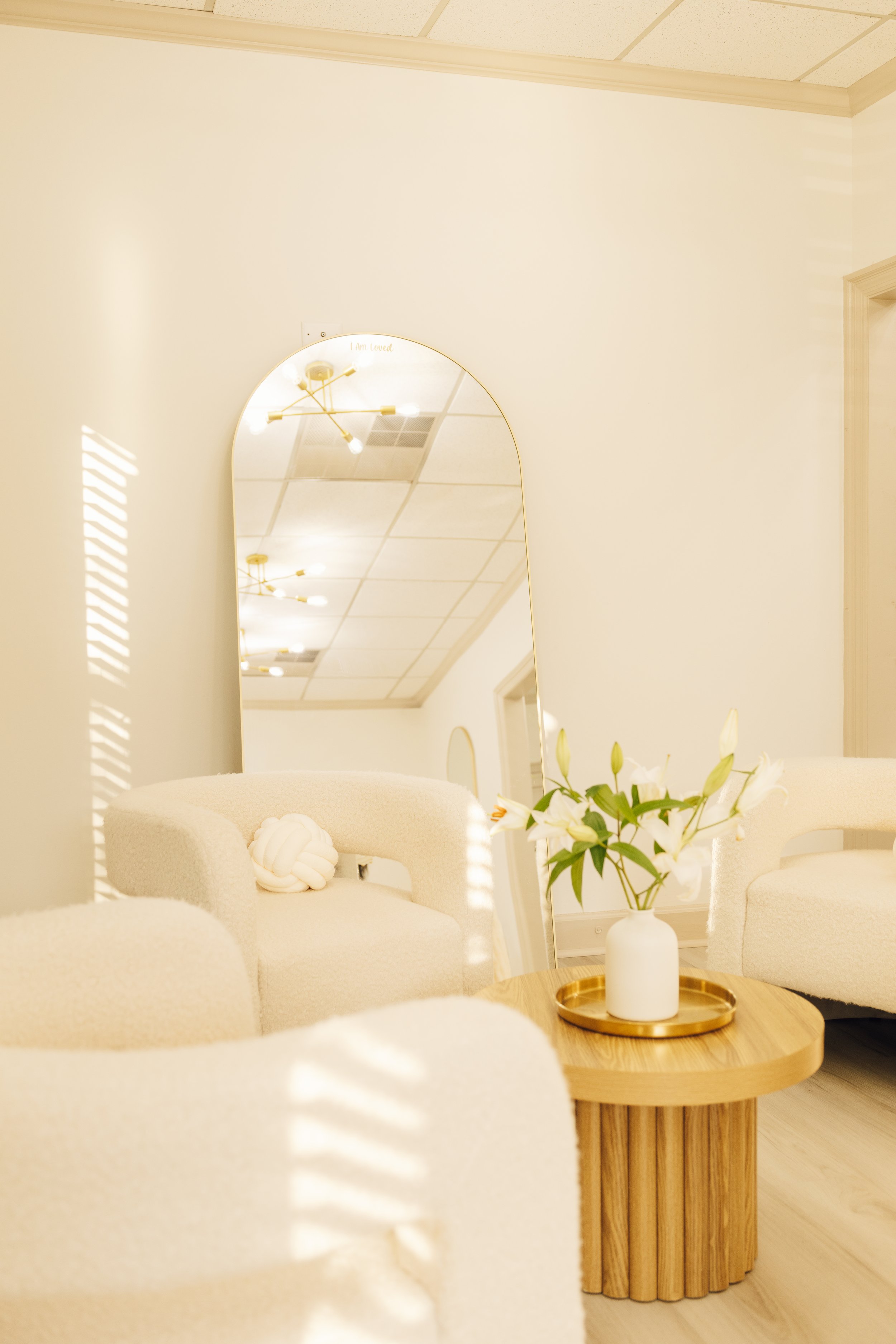 A cozy, minimalistic sitting area with cream-colored armchairs, a round wooden table with a white vase of white flowers, a large mirror on the wall, and a ceiling light fixture reflected in the mirror.