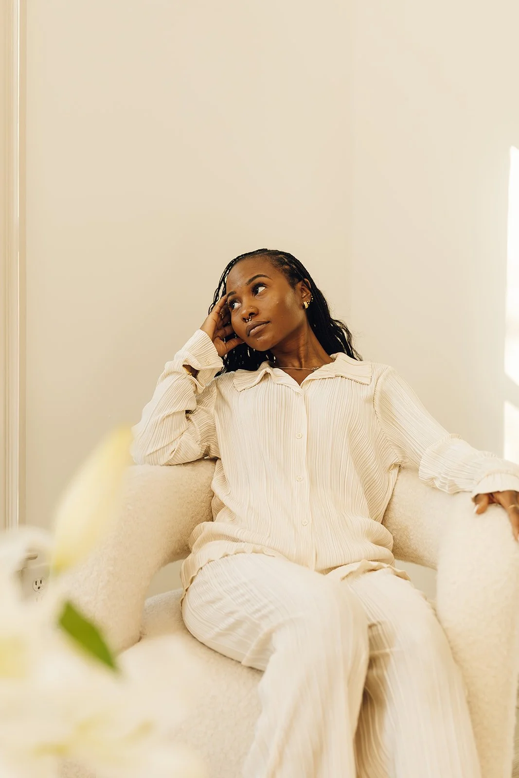 A woman with dark skin and braided hair sitting in a cream-colored armchair, wearing light cream-colored pajamas, resting her head on her hand, and looking thoughtfully to the side in a well-lit room.