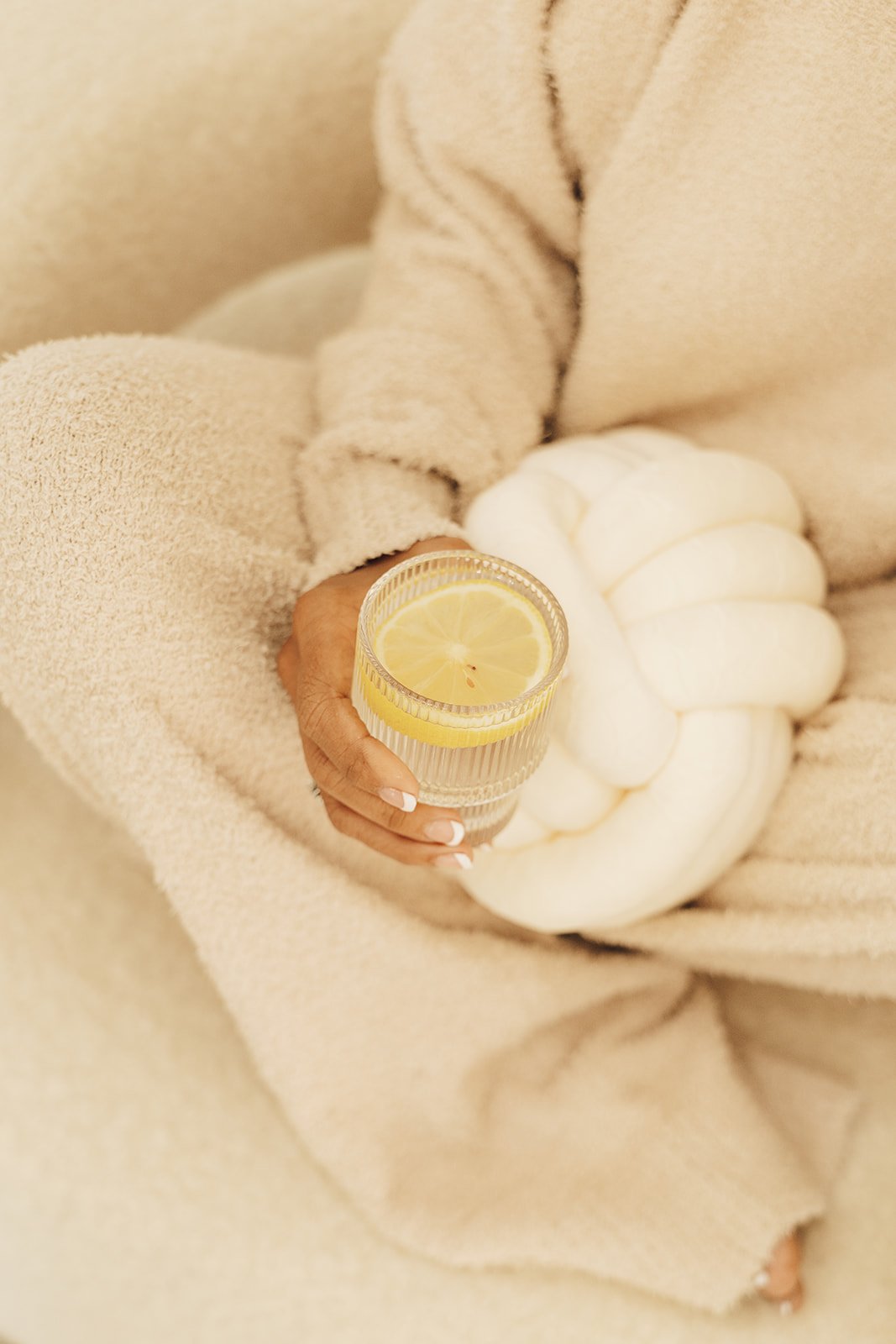 A person sitting on a beige couch holds a glass of water with lemon slices. They are wearing a beige, textured, long-sleeved sweater and have a white, plush, pumpkin-shaped cushion on their lap.