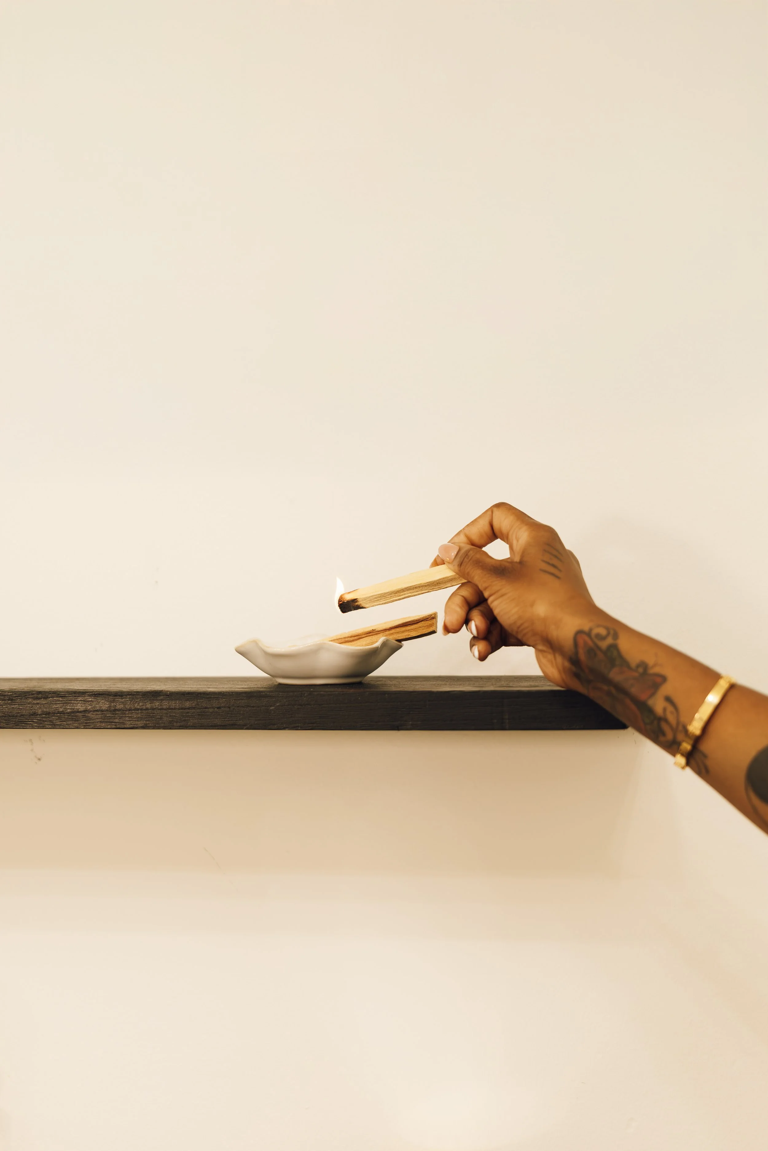 A person with tattoos and a gold bracelet holds a matchstick with a lit match, igniting incense in a white dish on a black shelf against a plain white wall.
