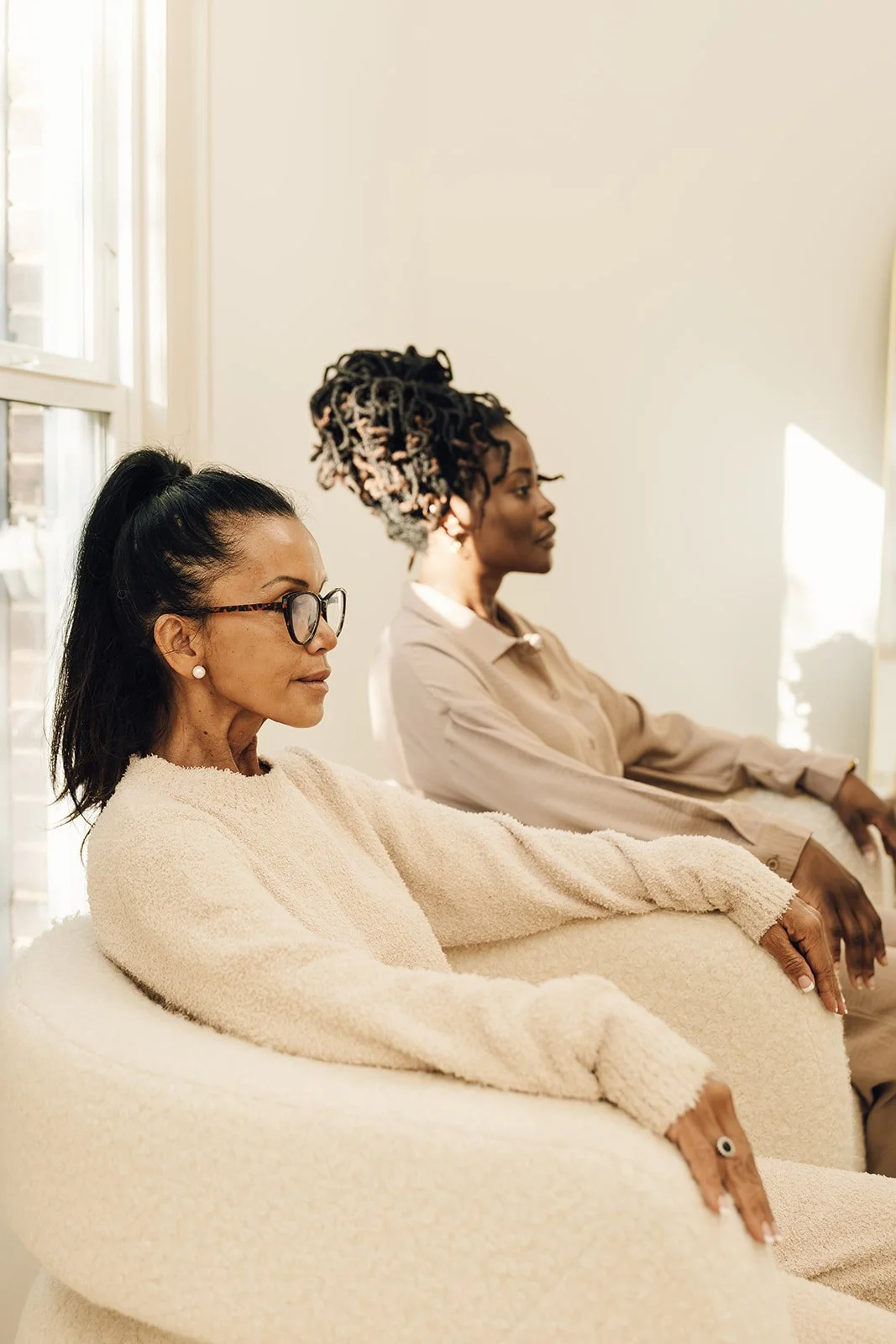 Two women sitting on cream-colored sofas in a bright room with sunlight streaming through windows, both with a calm, focused expression.