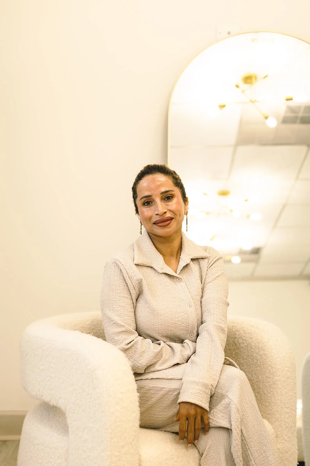 A woman in a beige outfit sits on a plush white chair in a room with cream-colored walls and a large mirror reflecting ceiling lights.