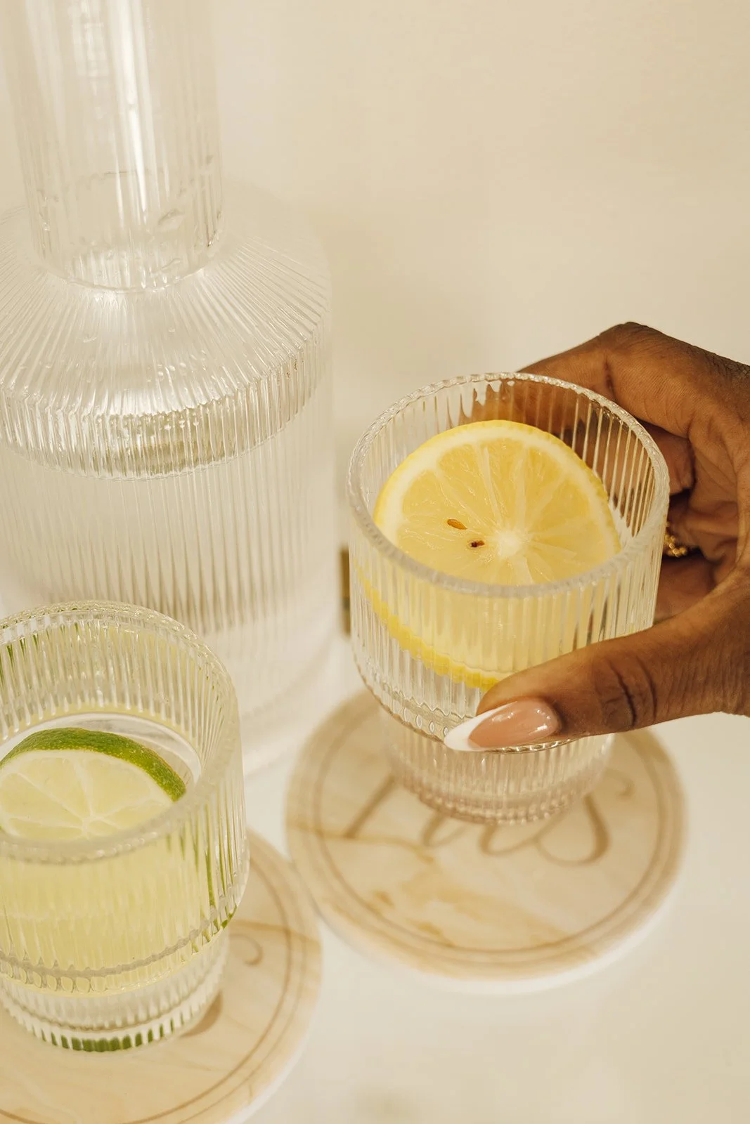 A person's hand holding a glass with a lemon slice, next to a glass with a lime slice, both on round wooden coasters, with a large glass water dispenser in the background.
