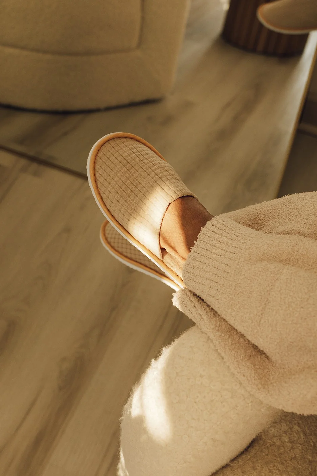 Person wearing beige slippers and cream-colored cozy pants, sitting on a wooden floor next to a brown upholstered chair.