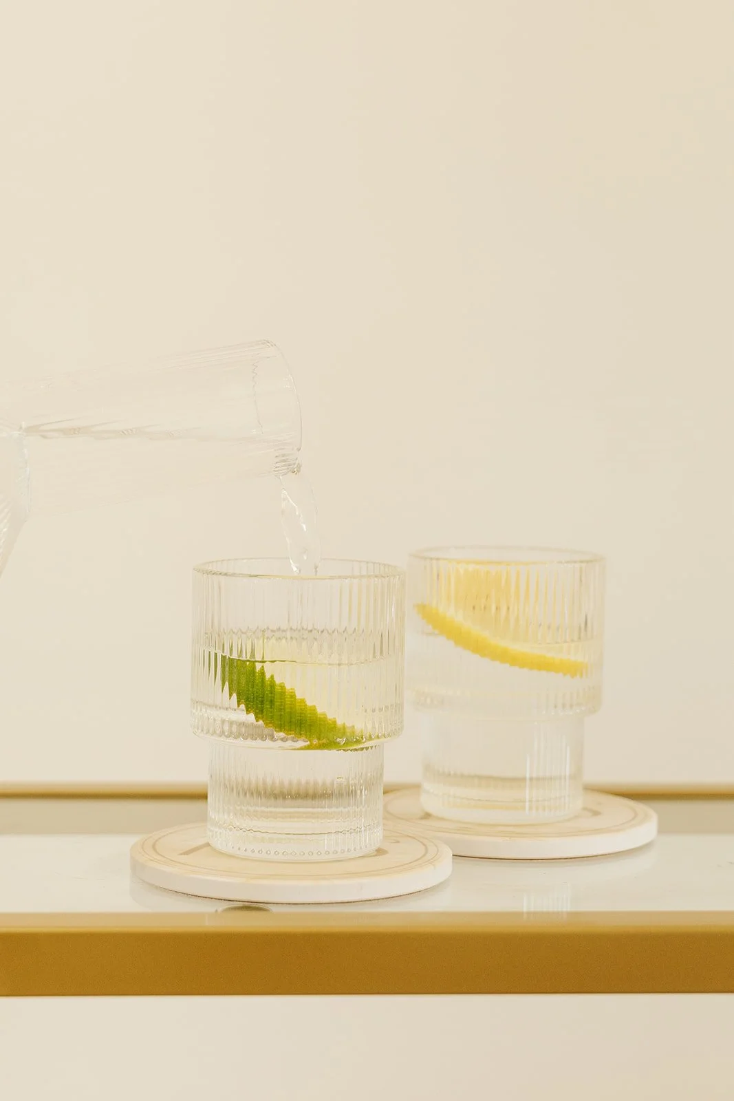 A glass of water with a lime wedge inside, with water being poured from a pitcher, next to another glass with a lemon wedge, both on round coasters on a white surface.