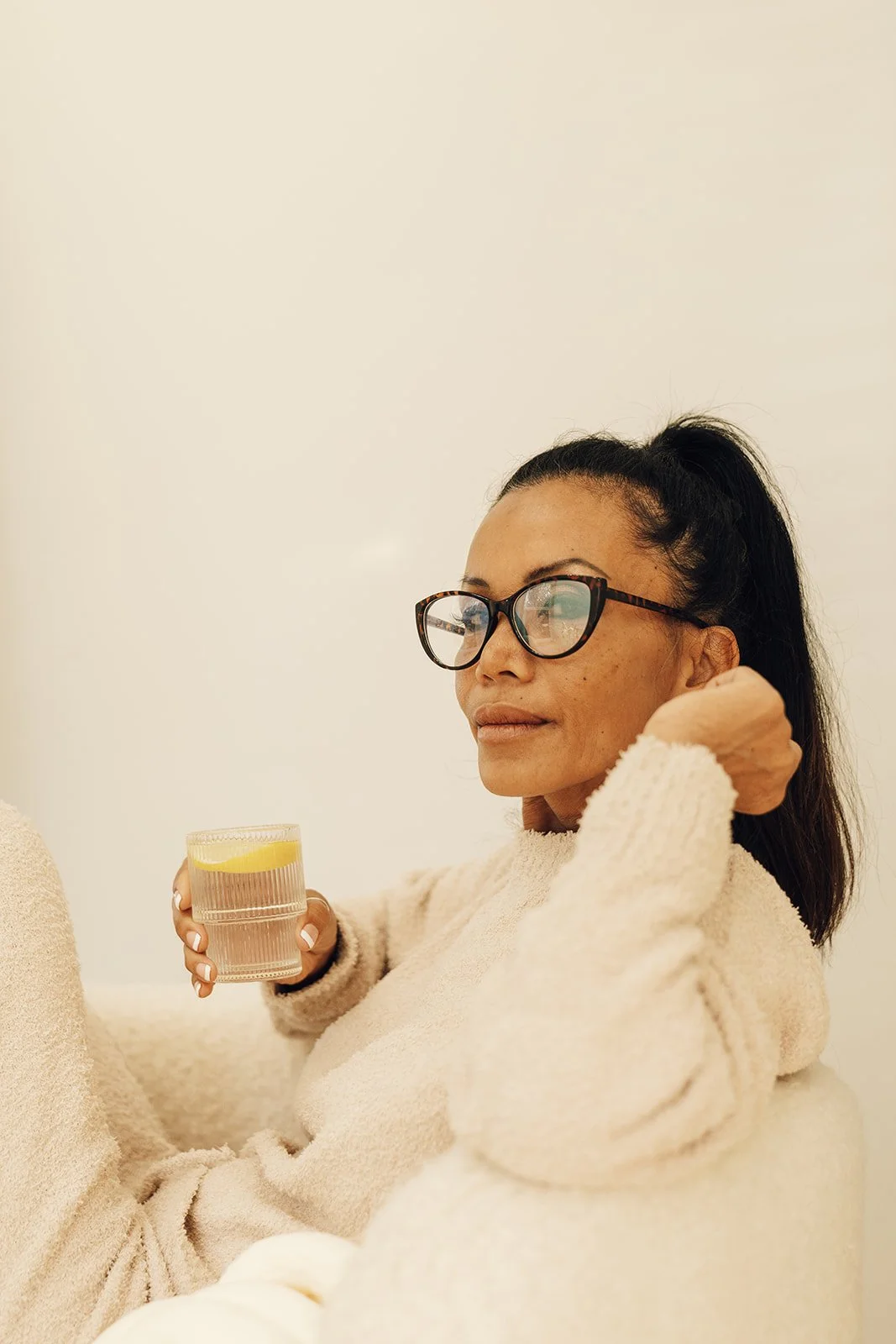 Woman with glasses relaxing on a cream-colored couch, holding a glass of water with lemon, in a minimalistic room with plain white walls.