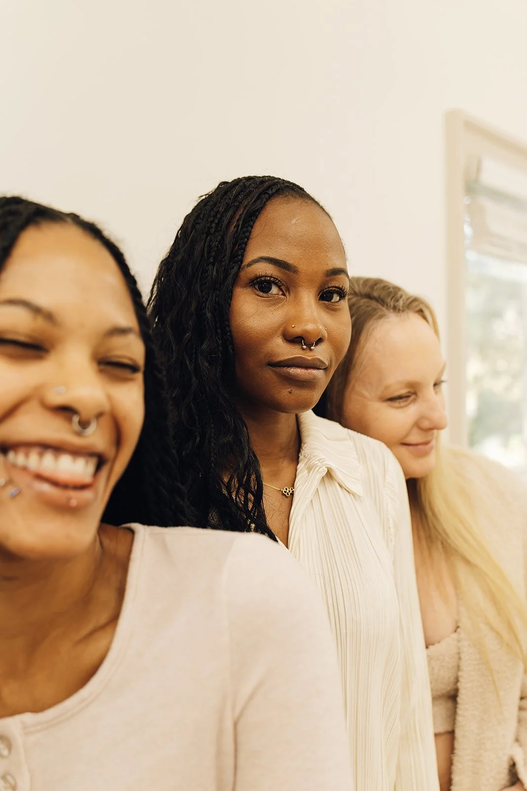 Three women standing indoors near a window, smiling and looking at the camera.