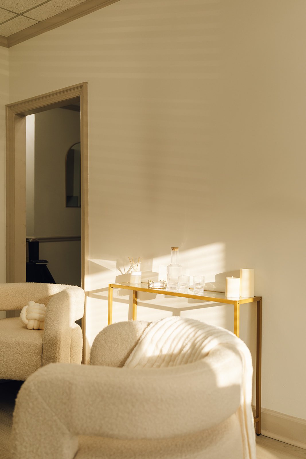Sunlight streaming onto a white sofa and a minimalistic gold console table with candles, glassware, and a decanter in a cozy, well-lit room.