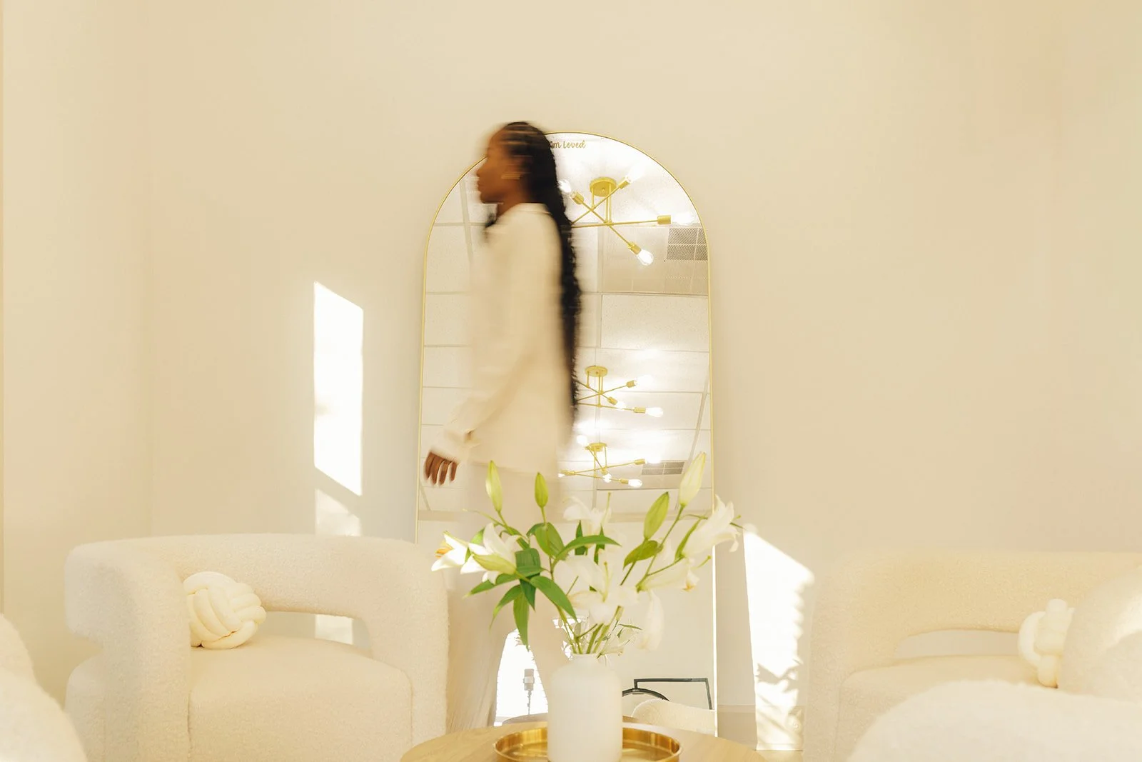 Interior with a white sofa, a white armchair, a round wooden table with a white vase of white lilies, a large mirror reflecting a woman with long dark hair, and warm lighting fixtures.