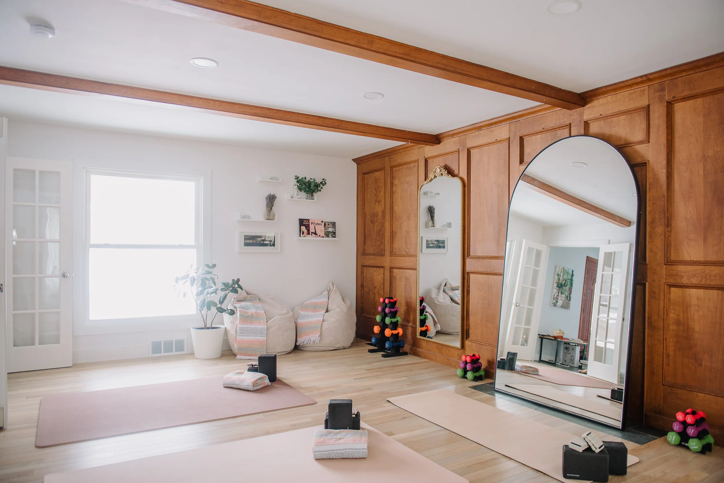 A bright home gym with workout mats, dumbbells, and exercise equipment, featuring a large mirror and a wooden wall with shelves and framed pictures.