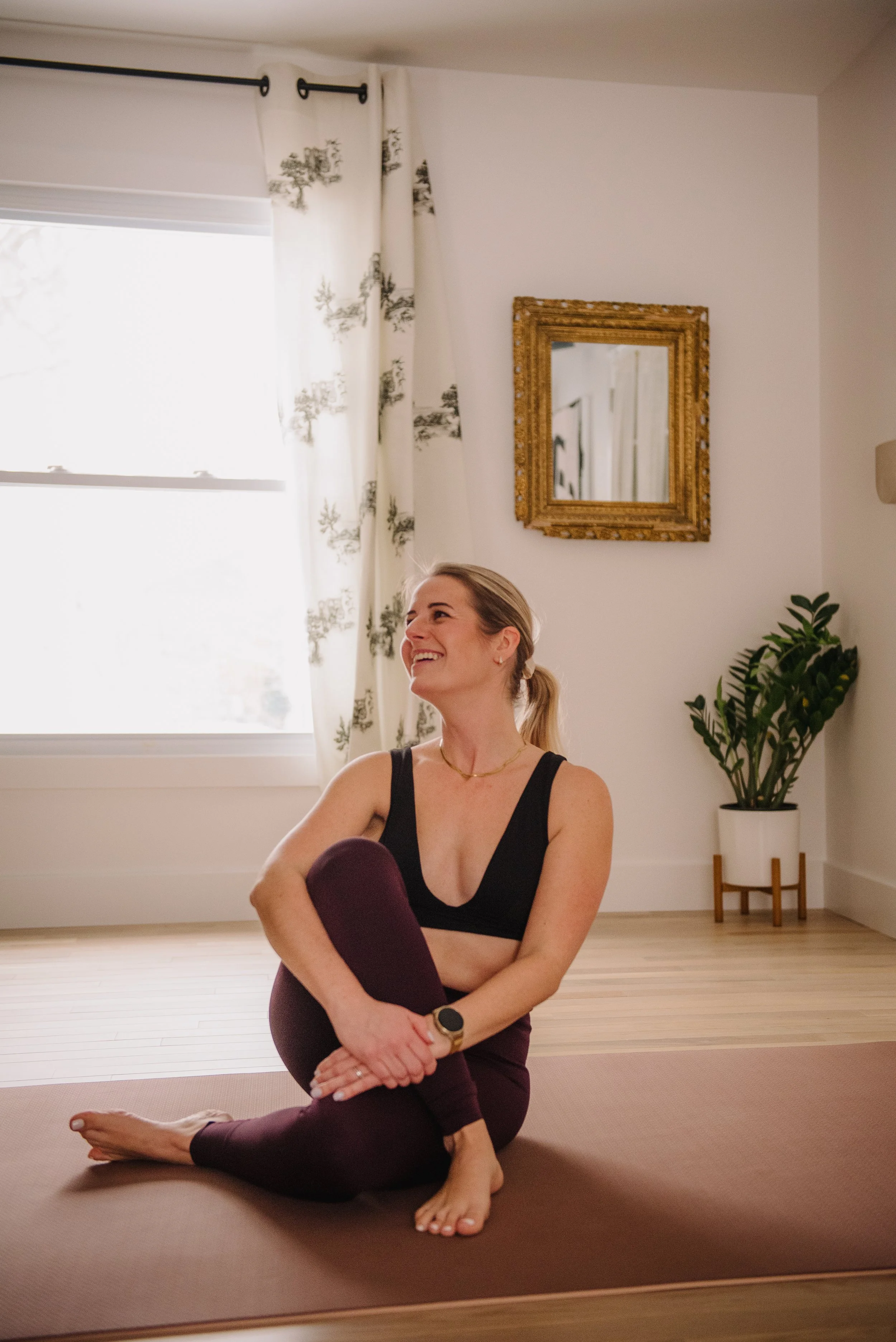 A woman practicing yoga indoors on a mat, smiling and looking relaxed, with natural light coming through a window with curtains.