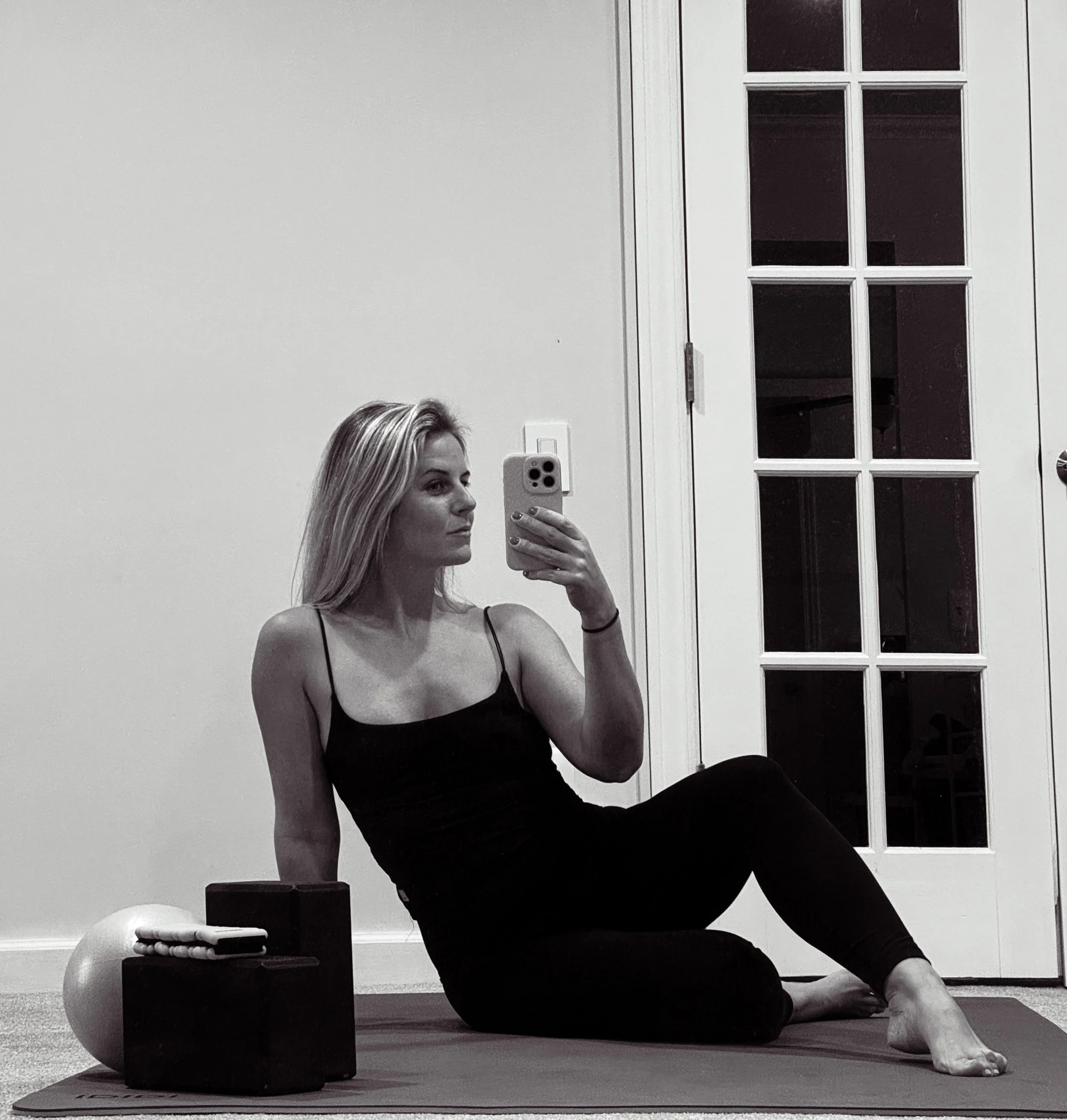 A woman with long blonde hair in a black tank top and leggings taking a mirror selfie with her smartphone while sitting on a yoga mat in a room. Exercise equipment, including a stability ball and yoga blocks, are nearby.
