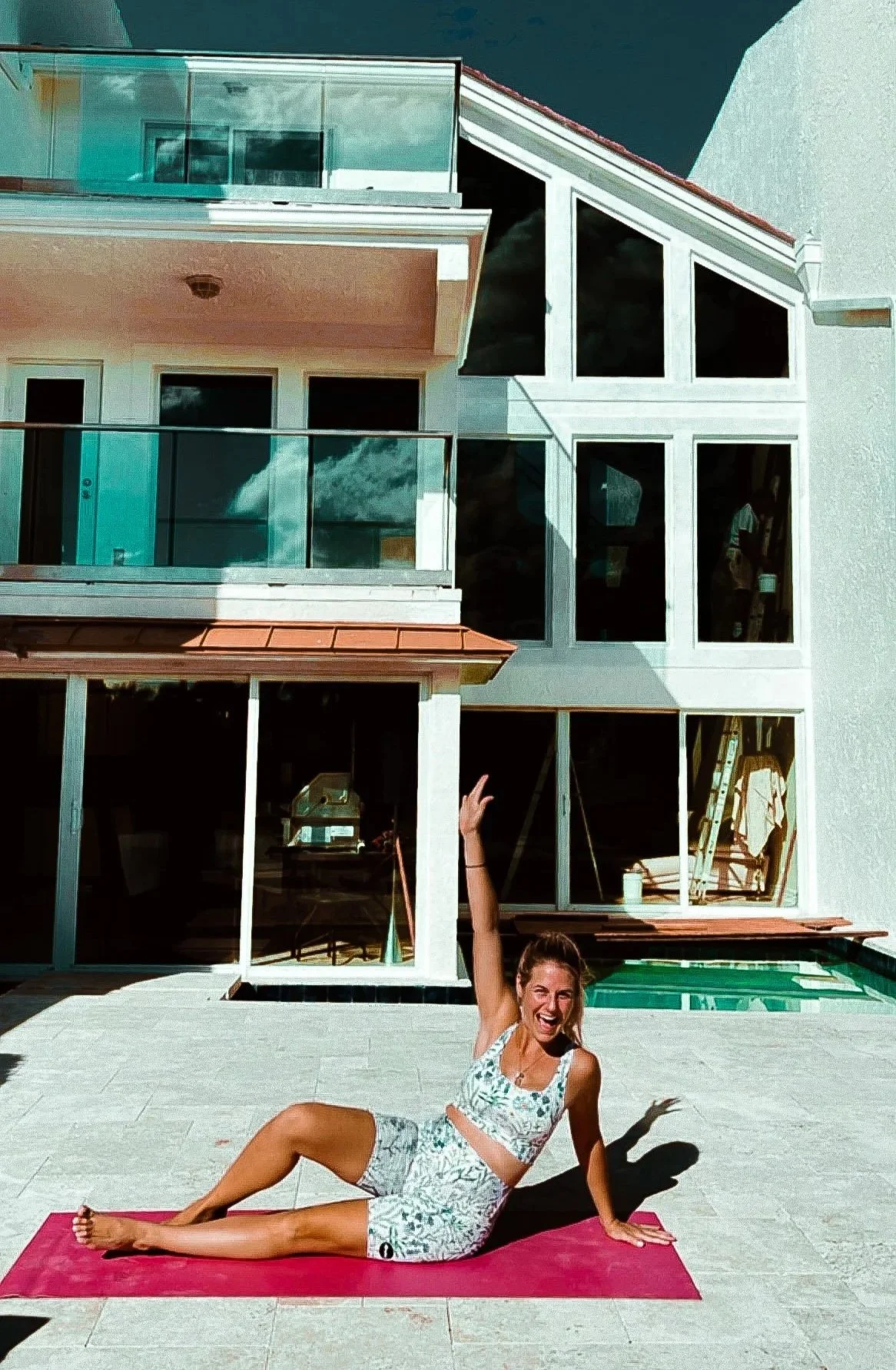 A woman lying on a pink yoga mat outdoors in front of a modern house with large glass windows and a balcony, raising her right hand and smiling.