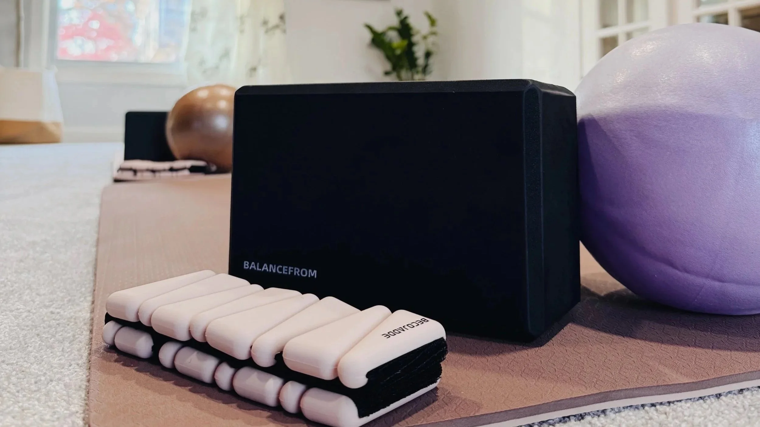 Yoga props including a black BalanceFrom yoga block, a pink and black balance pad, and a purple exercise ball on a yoga mat in a bright room with a window and a plant.