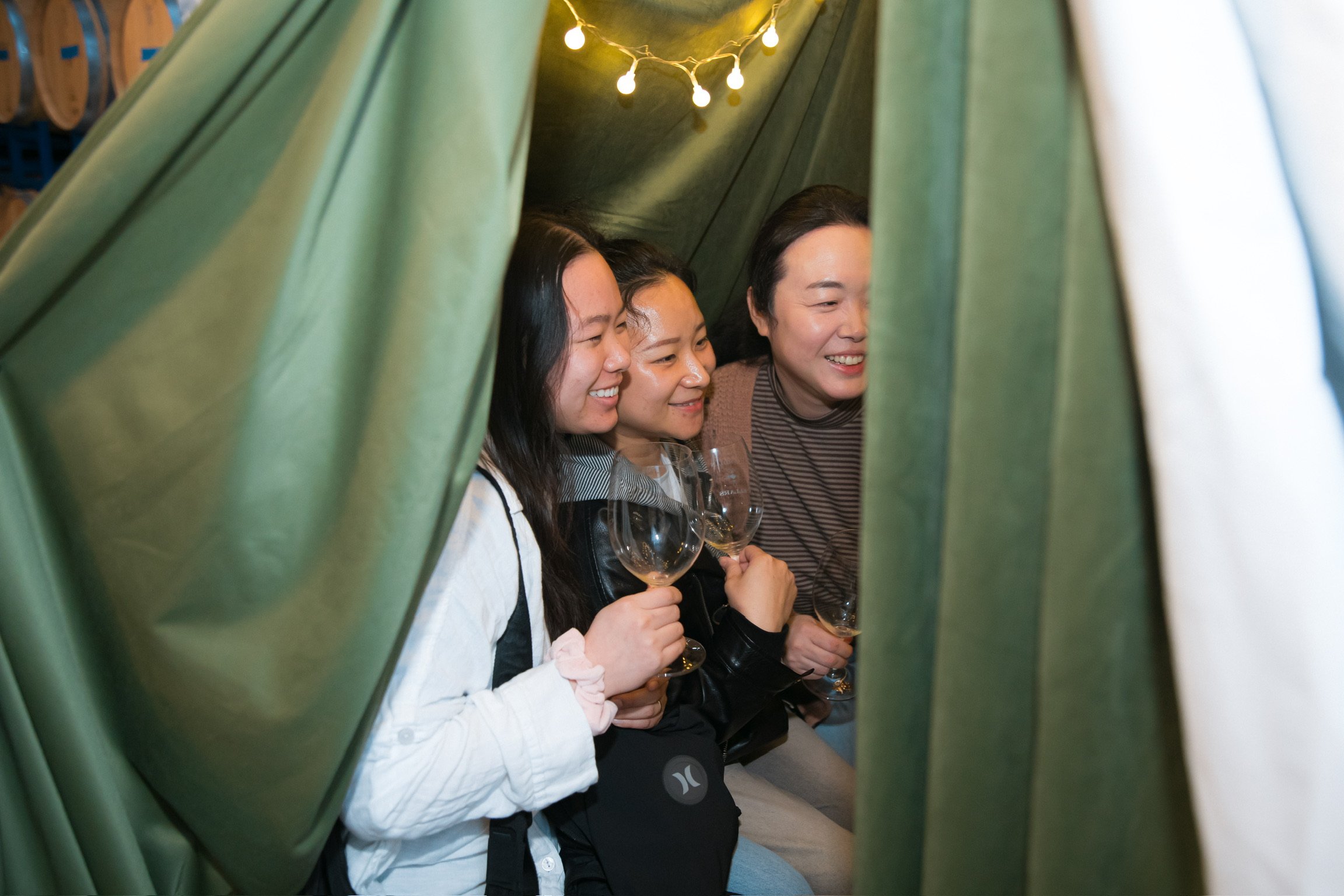 Three women smiling and holding wine glasses, sitting inside a cozy, green tent with string lights overhead.