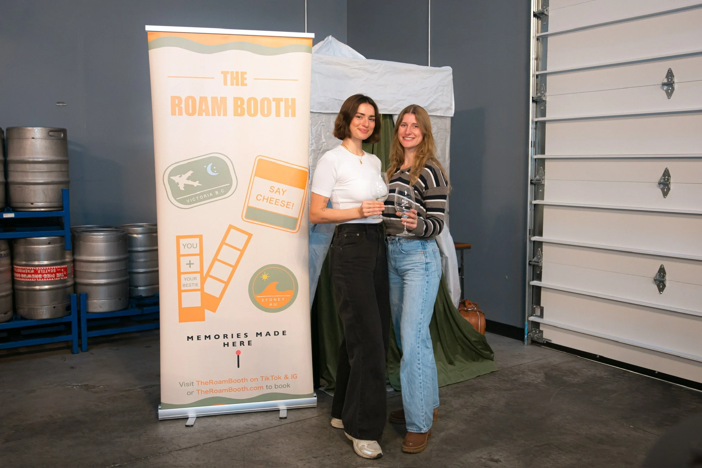 Two women standing next to each other at a photo booth, holding glasses, smiling. Behind them is a banner that reads 'The Roam Booth' with travel-themed icons and text.