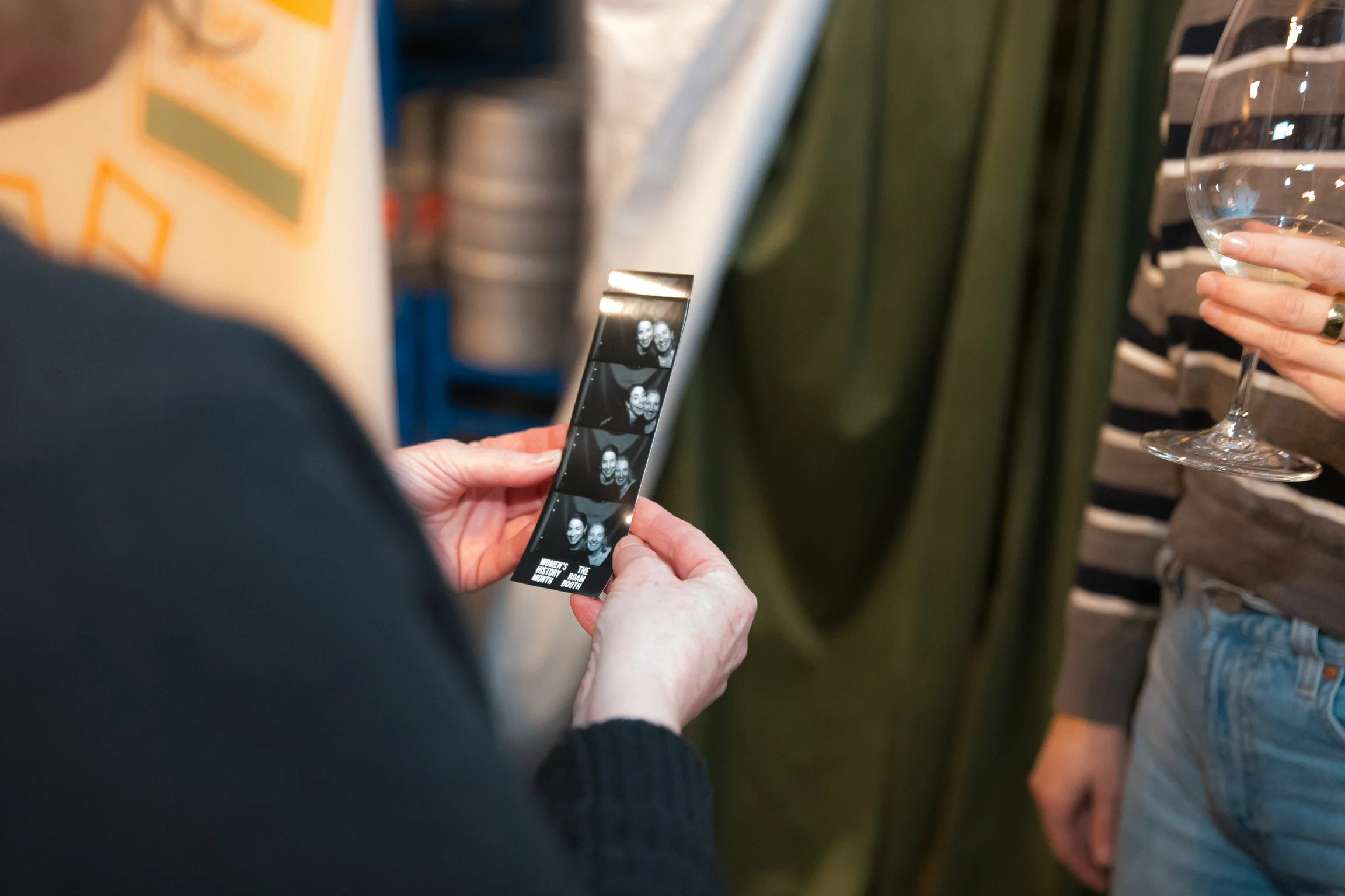 Person holding a photo strip with four images, showing two women smiling, in an indoor setting along with another person holding a wine glass.