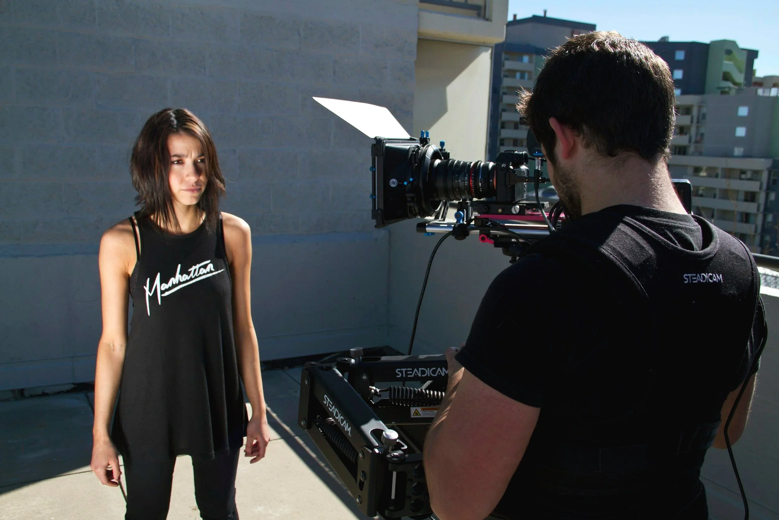 A woman in a black tank top with 'Manhattan' written on it poses for a photo on a rooftop with city buildings in the background. A man operating a professional camera on a stabilizer films her.