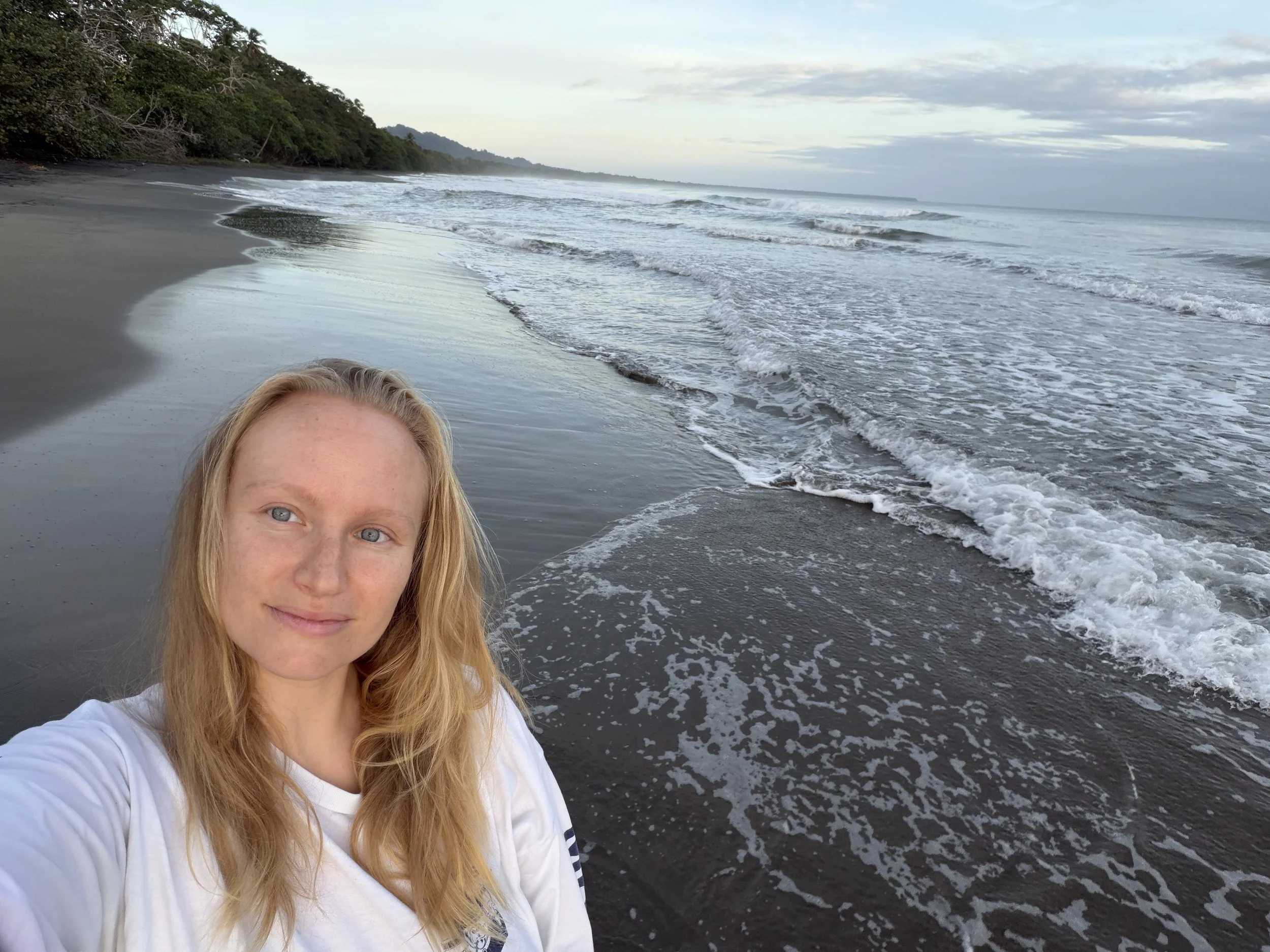 A woman with long blonde hair taking a selfie on a beach at sunset or sunrise, with the ocean waves and sandy shoreline in the background.