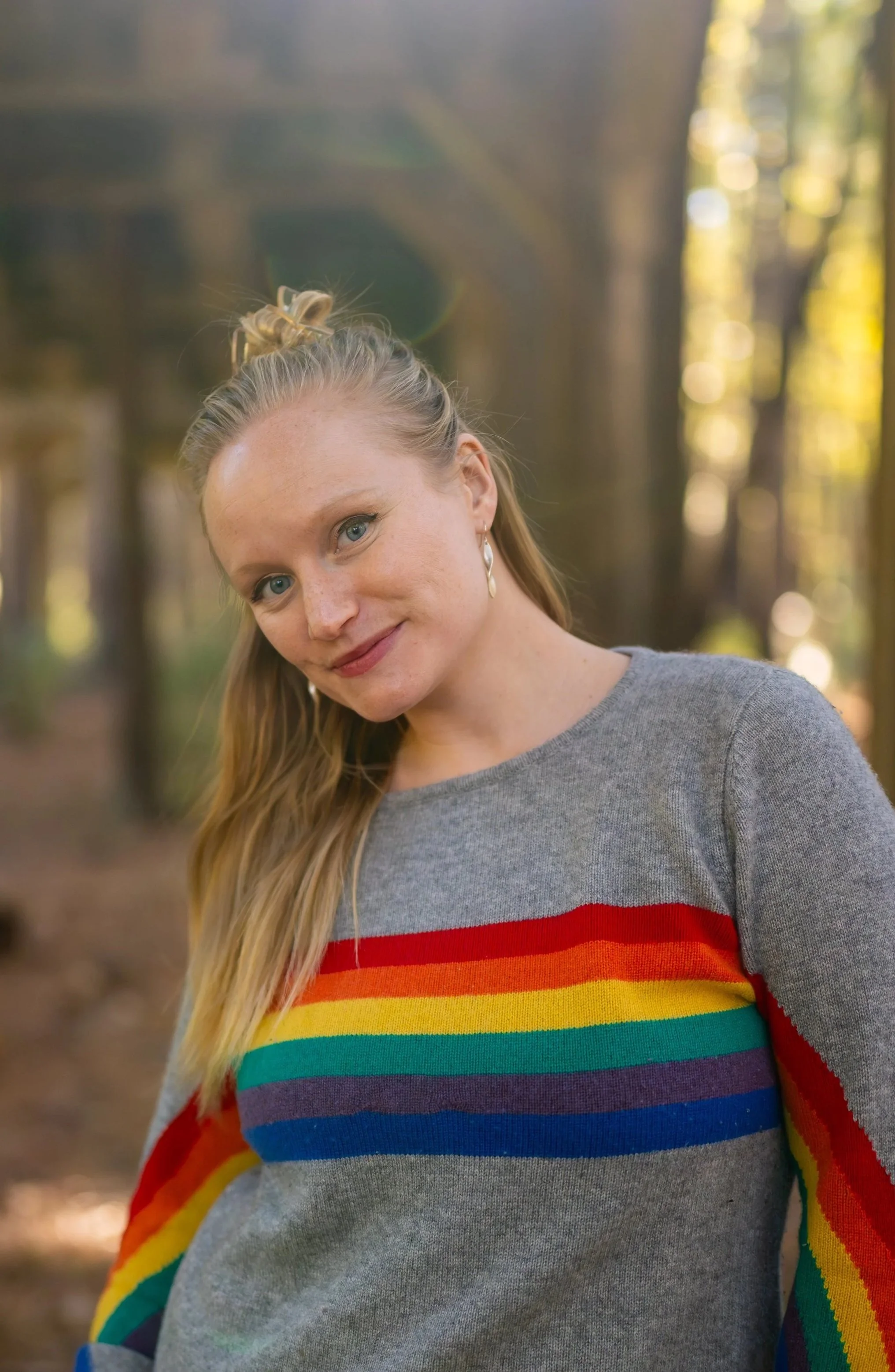 A woman with long blonde hair wearing a gray sweater with rainbow stripes, standing outdoors in a forest with blurred trees and sunlight in the background.