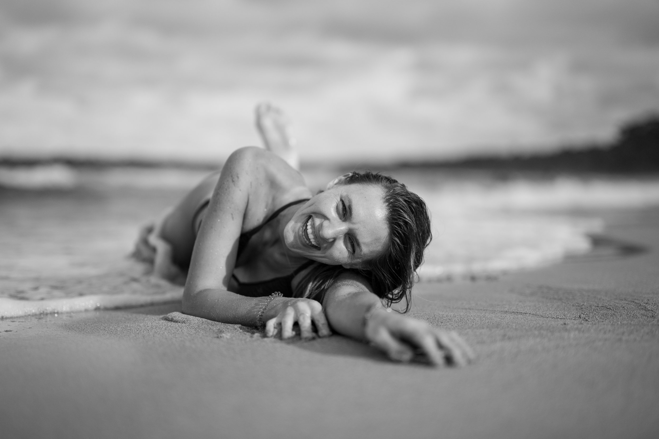 A woman lying on her side on the beach, smiling and reaching towards the camera, with the ocean and cloudy sky in the background.