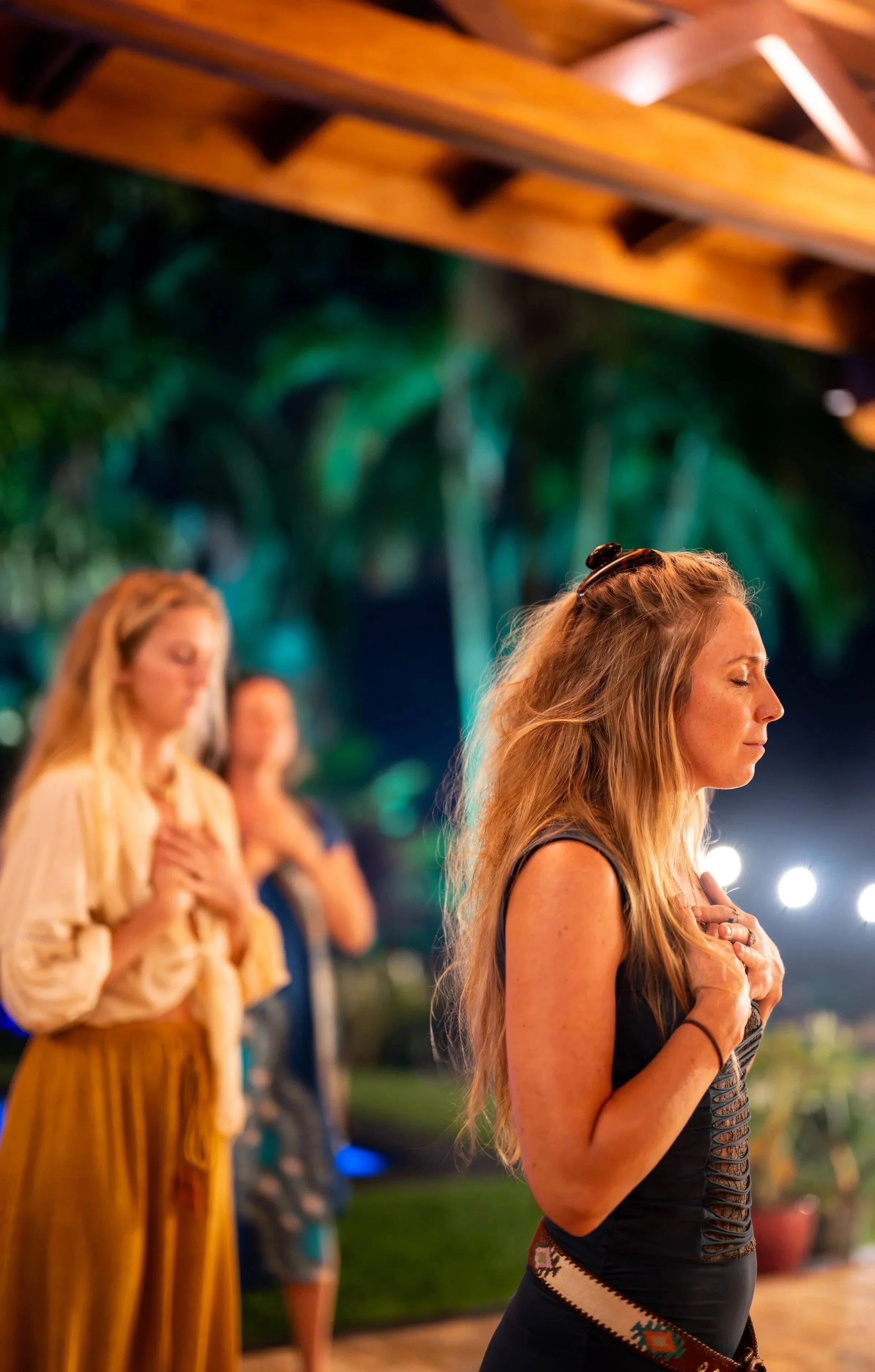 Three women standing outdoors at night with eyes closed and hands over their hearts, participating in a prayer or meditation. The woman in the foreground has long, wavy red hair and is wearing a black sleeveless top. Two women in the background are blurred, one with blonde hair and a beige cardigan, the other with darker hair and a dark blue top. The setting features an overhead wooden structure with string lights and a lush, green background.