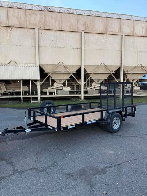 A black utility trailer with a mesh ramp at the back, parked on a paved surface in front of an industrial building with large beige tanks.