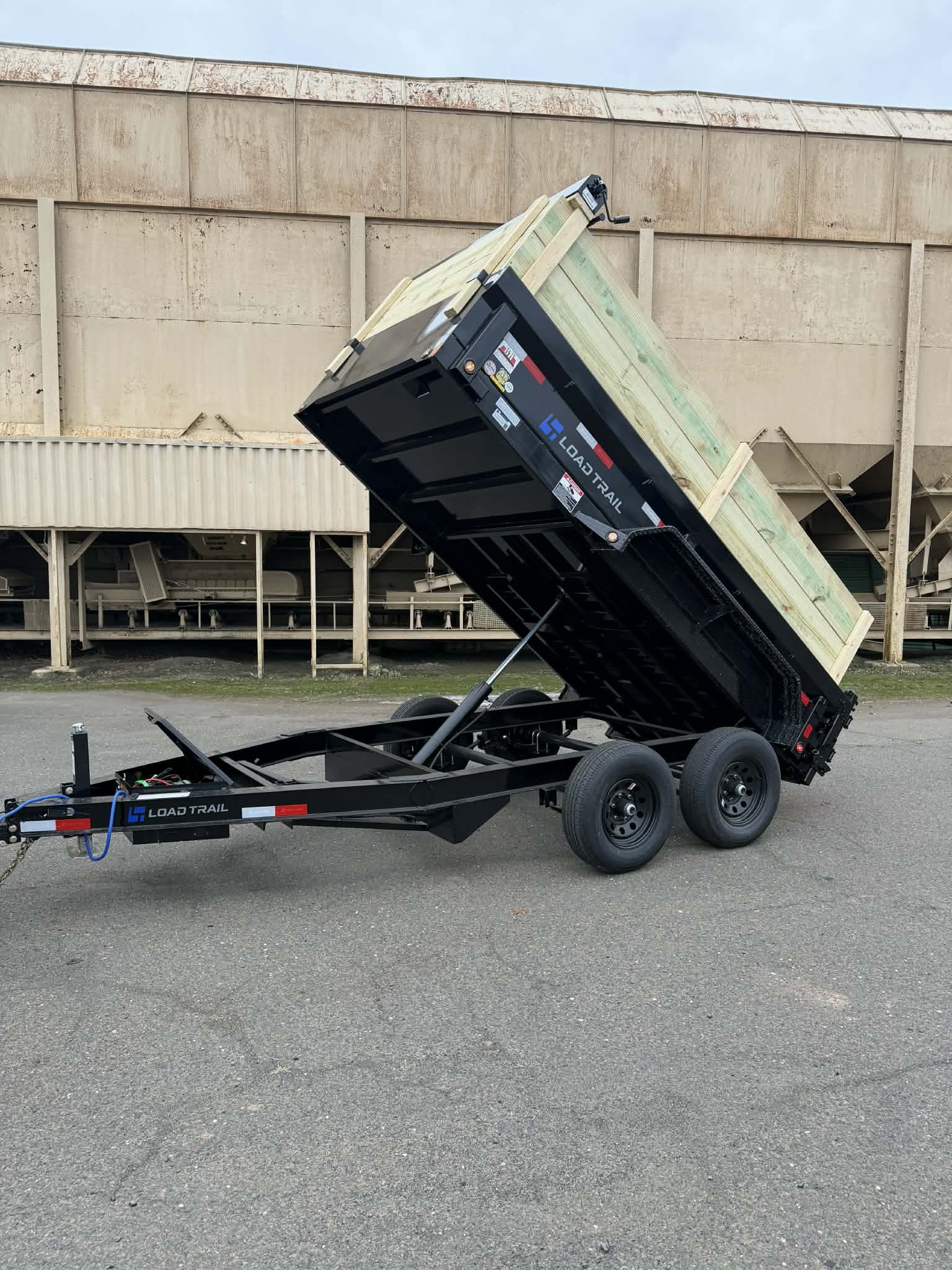 Black load trail dump trailer with raised bed, parked on asphalt in front of a beige wall.